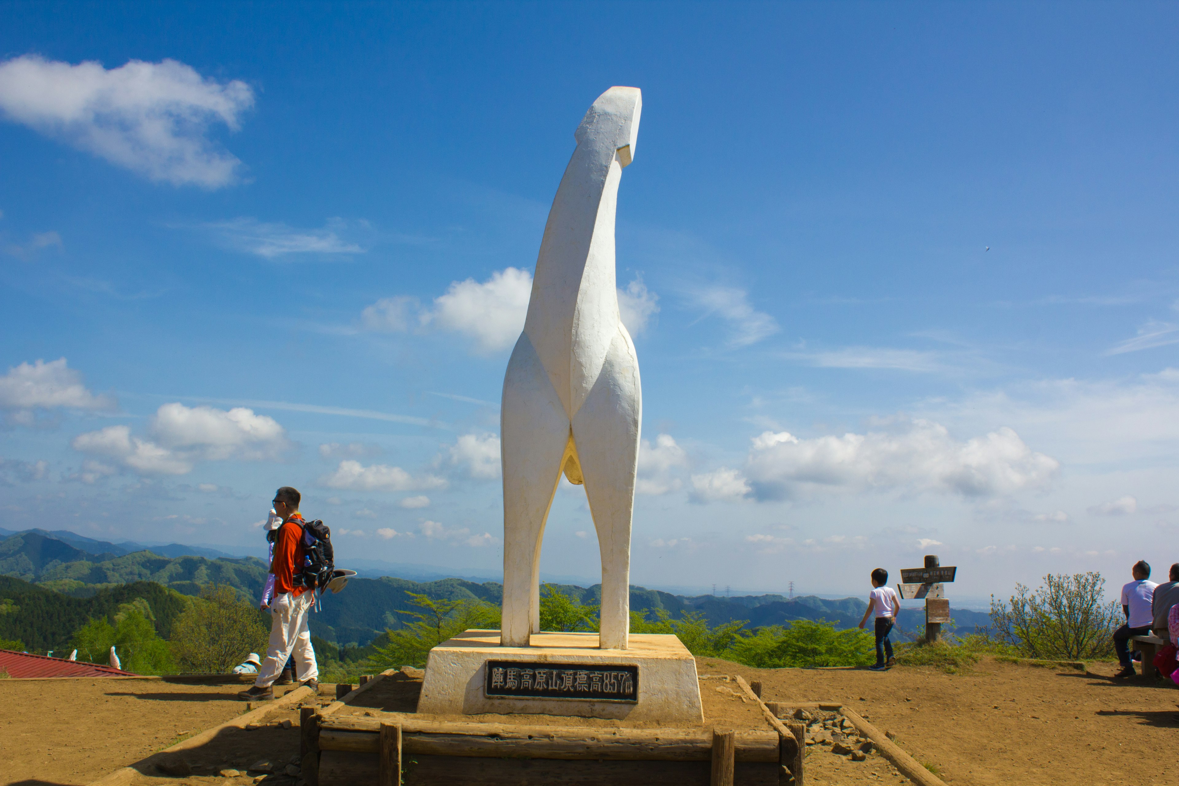 白い犬の像が立つ山の頂上に人々が集まる風景