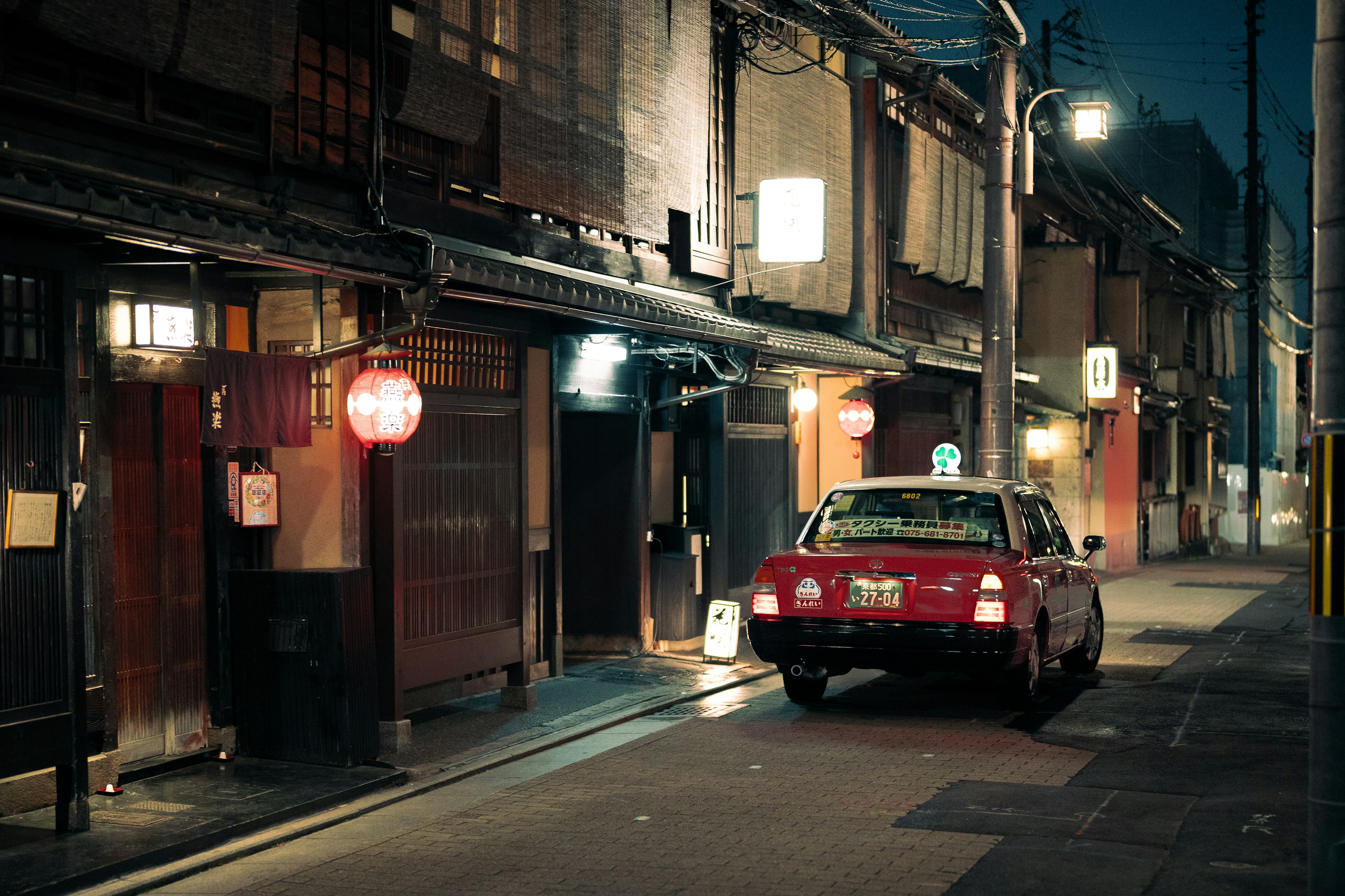 Ein rotes Taxi, das nachts in einer traditionellen Straße von Kyoto mit Laternen und beleuchteten Gebäuden parkt