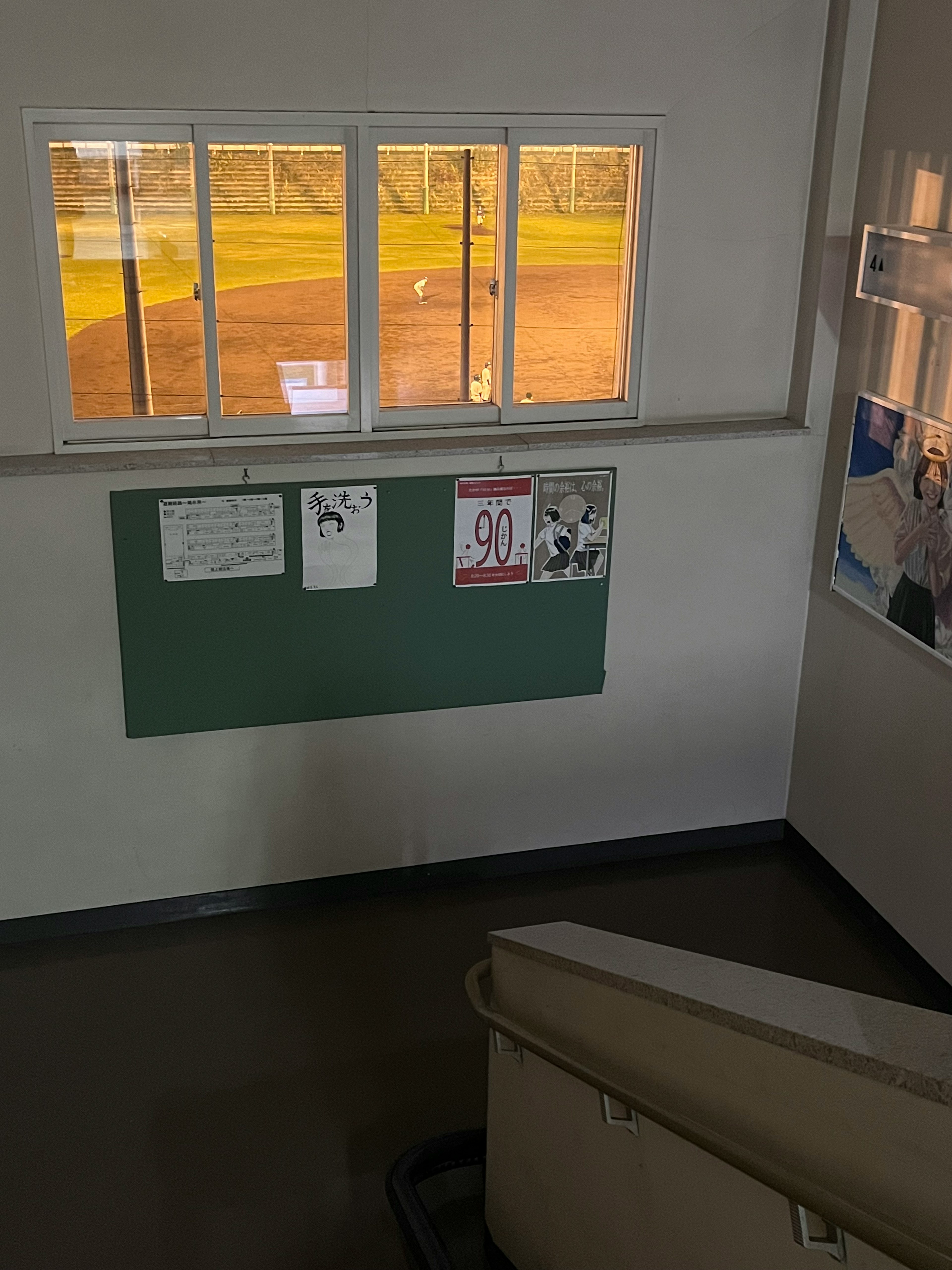 View of a baseball field through a window from a stairway with a bulletin board