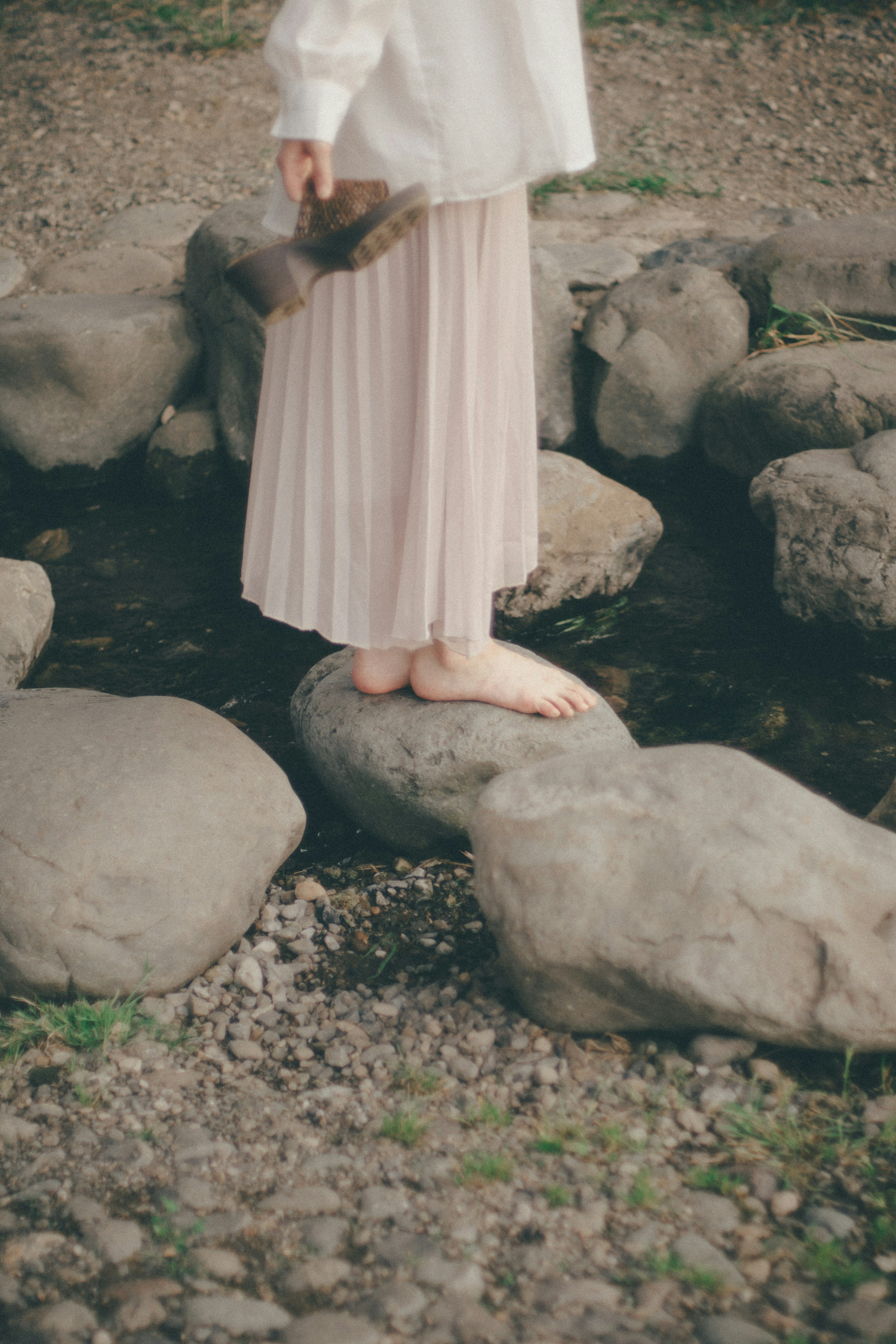 A woman standing on rocks by a stream with bare feet and a soft skirt