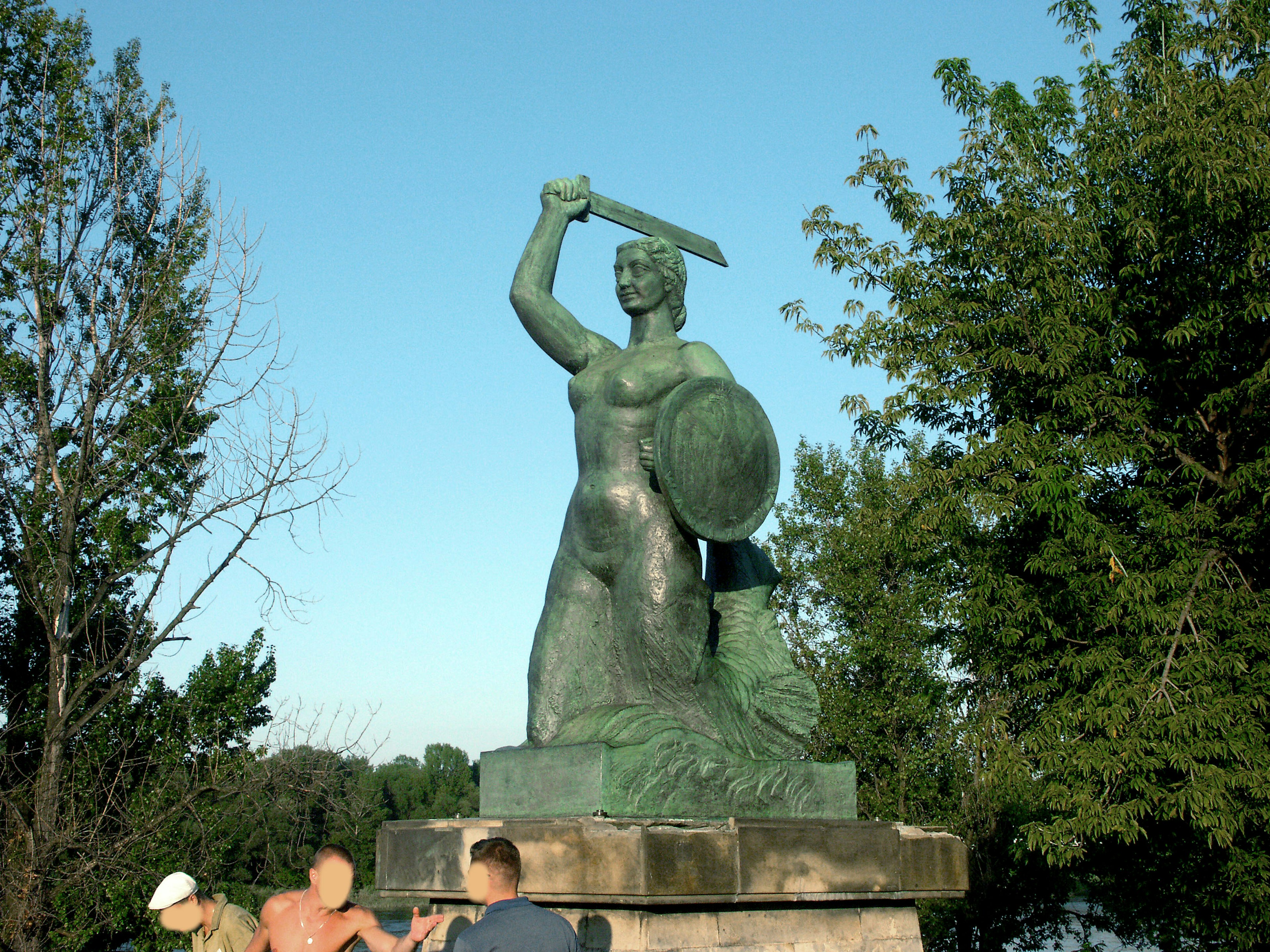 Eine Statue einer Frau, die unter einem blauen Himmel ein Schwert und ein Schild hält, mit Menschen, die sich um sie versammeln