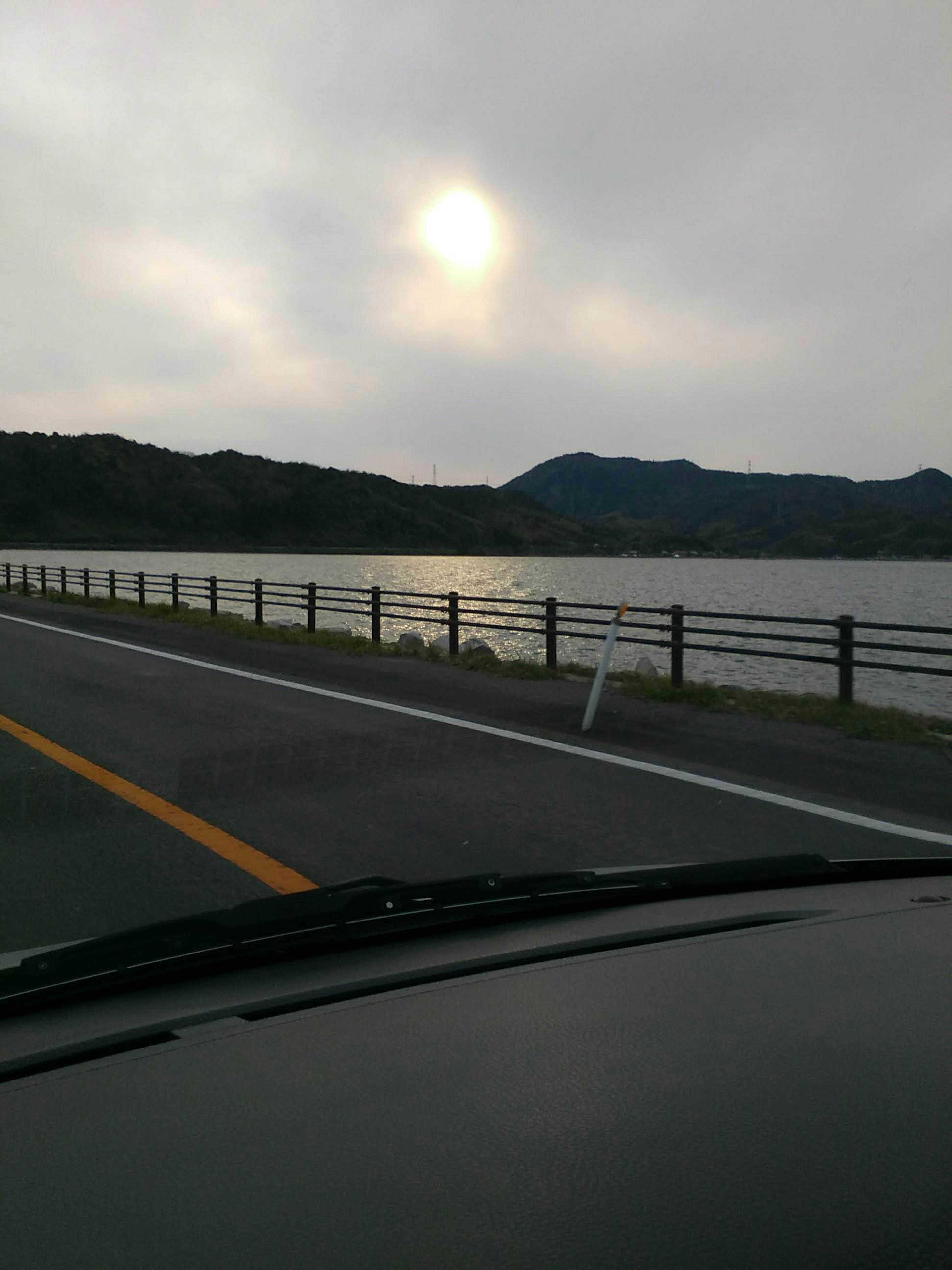 曇り空の下に広がる湖と山々の景色で、道路の横にはフェンスがあり、車のダッシュボードが見える