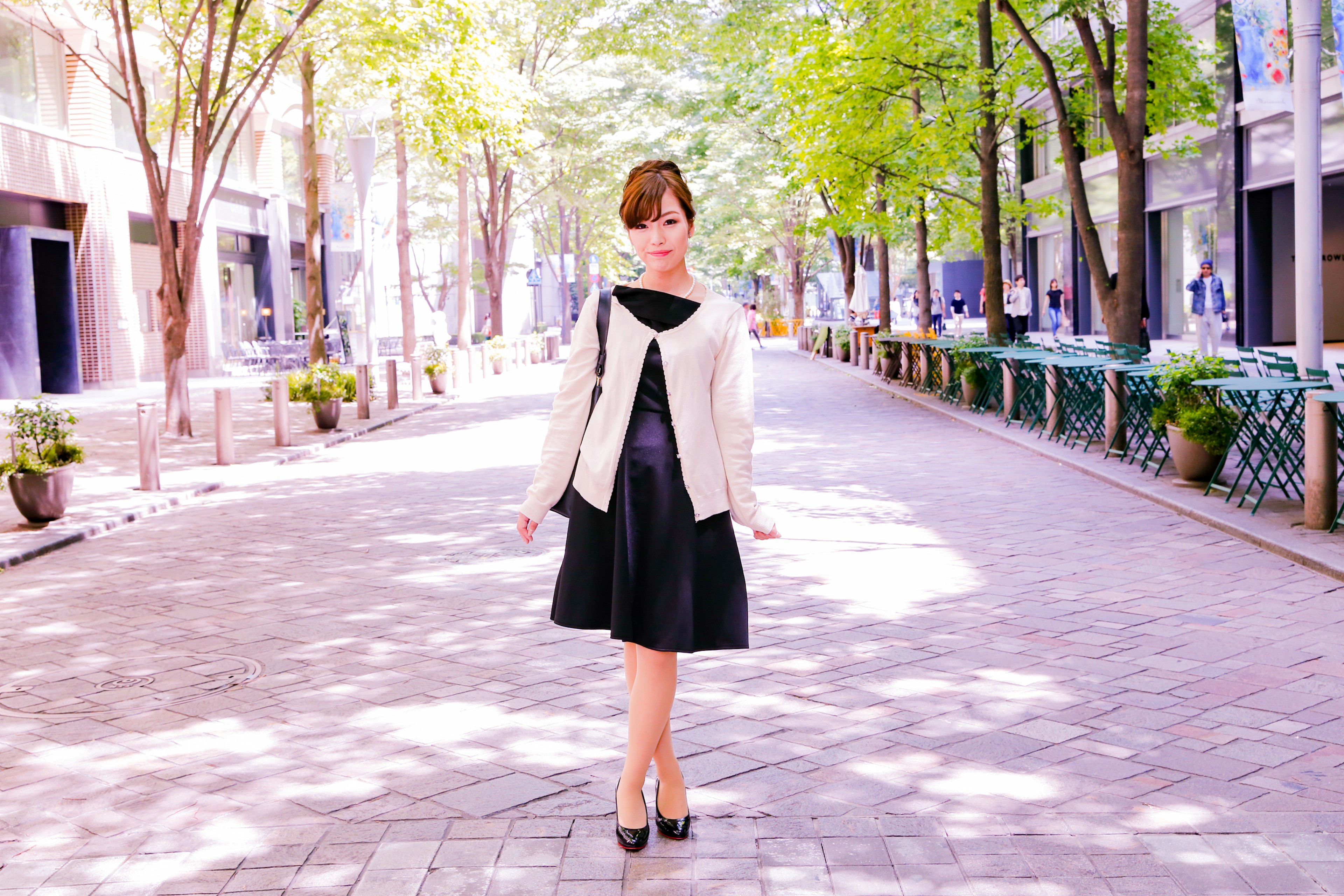 A woman posing from behind on a street, wearing a white jacket and black dress, surrounded by green trees