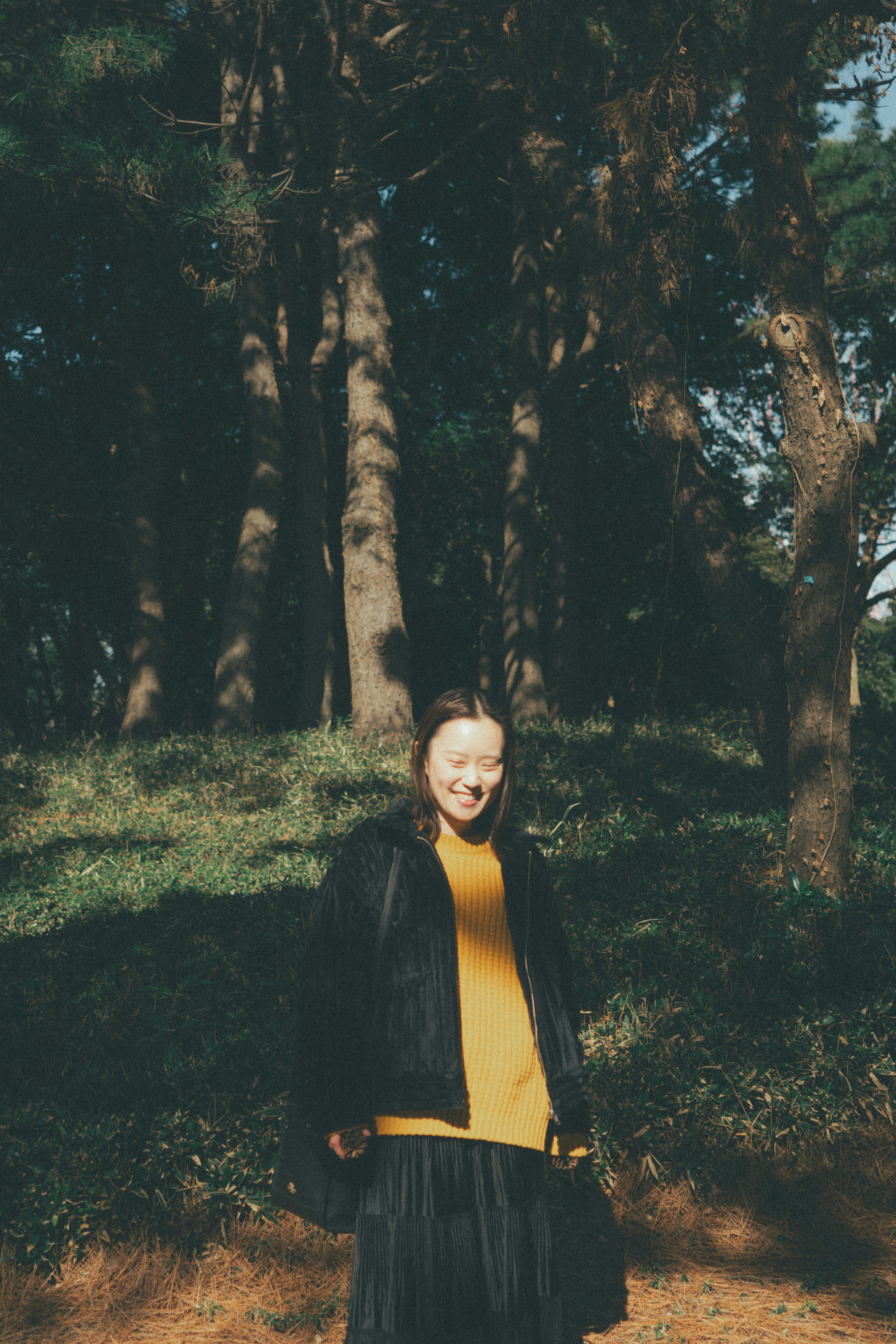 Foto einer lächelnden Frau im Wald, die einen leuchtend gelben Pullover und eine schwarze Jacke trägt