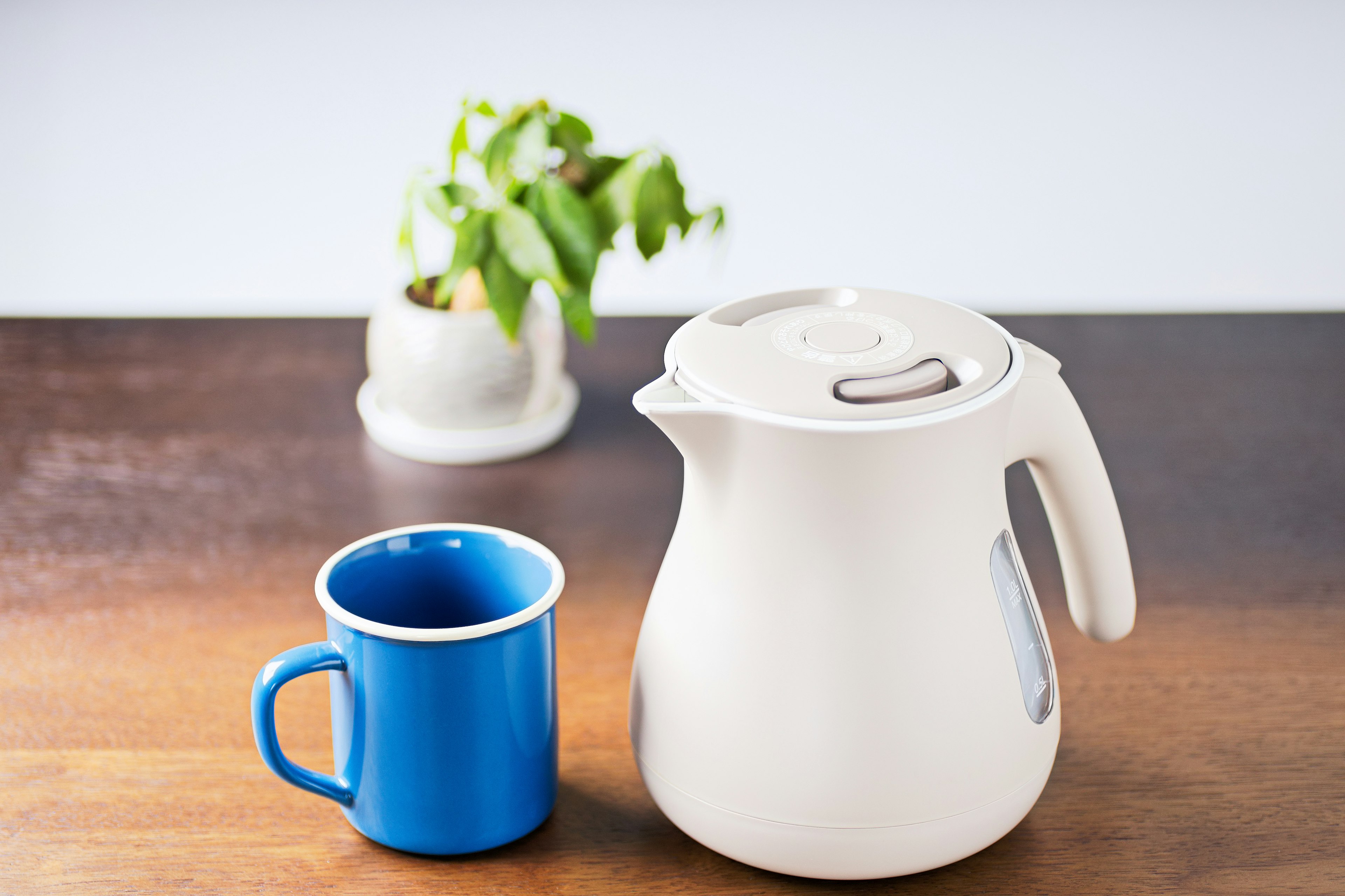 A white kettle and a blue mug placed on a wooden table with a plant in the background