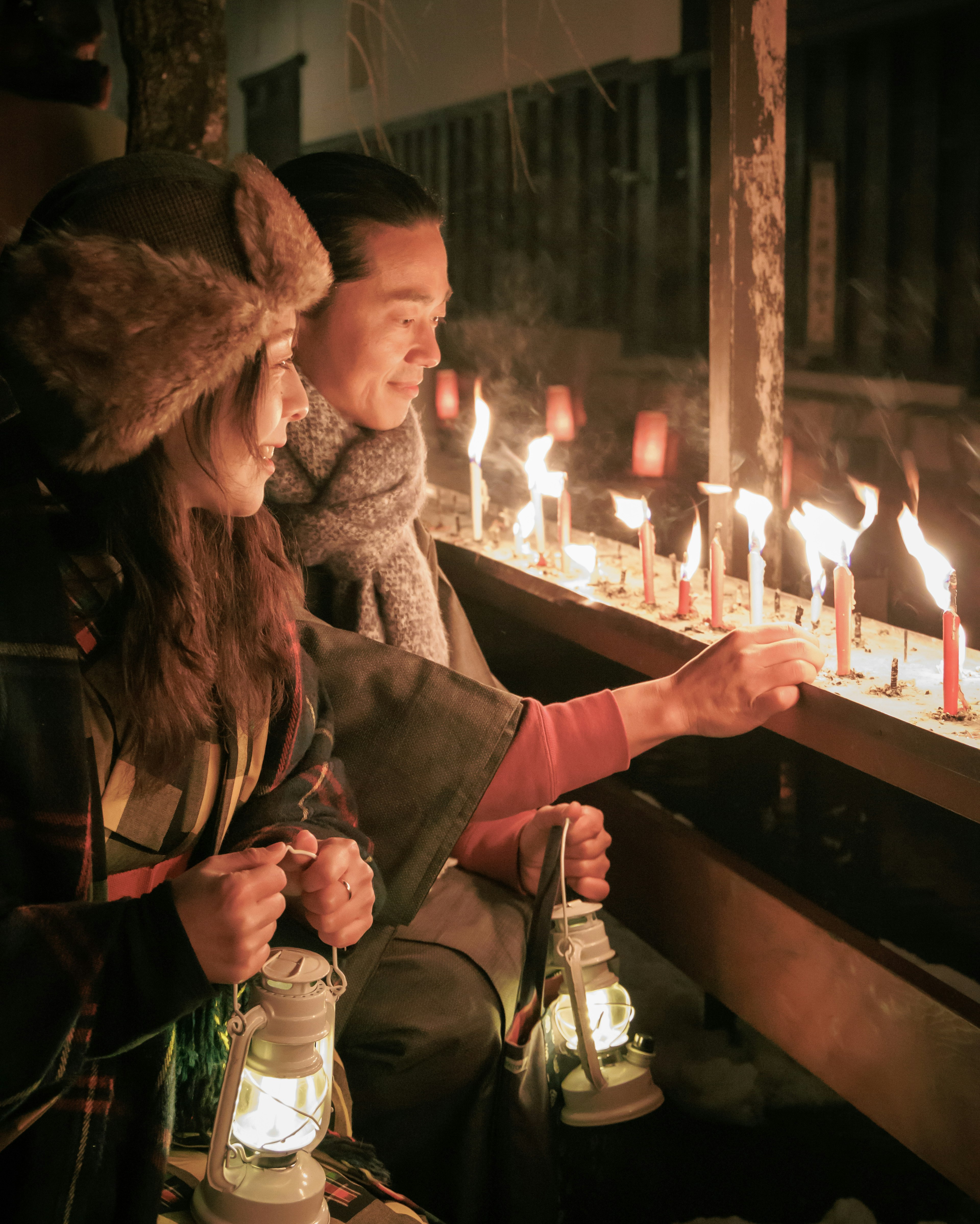 Two individuals seated in a dimly lit space surrounded by candlelight holding lanterns