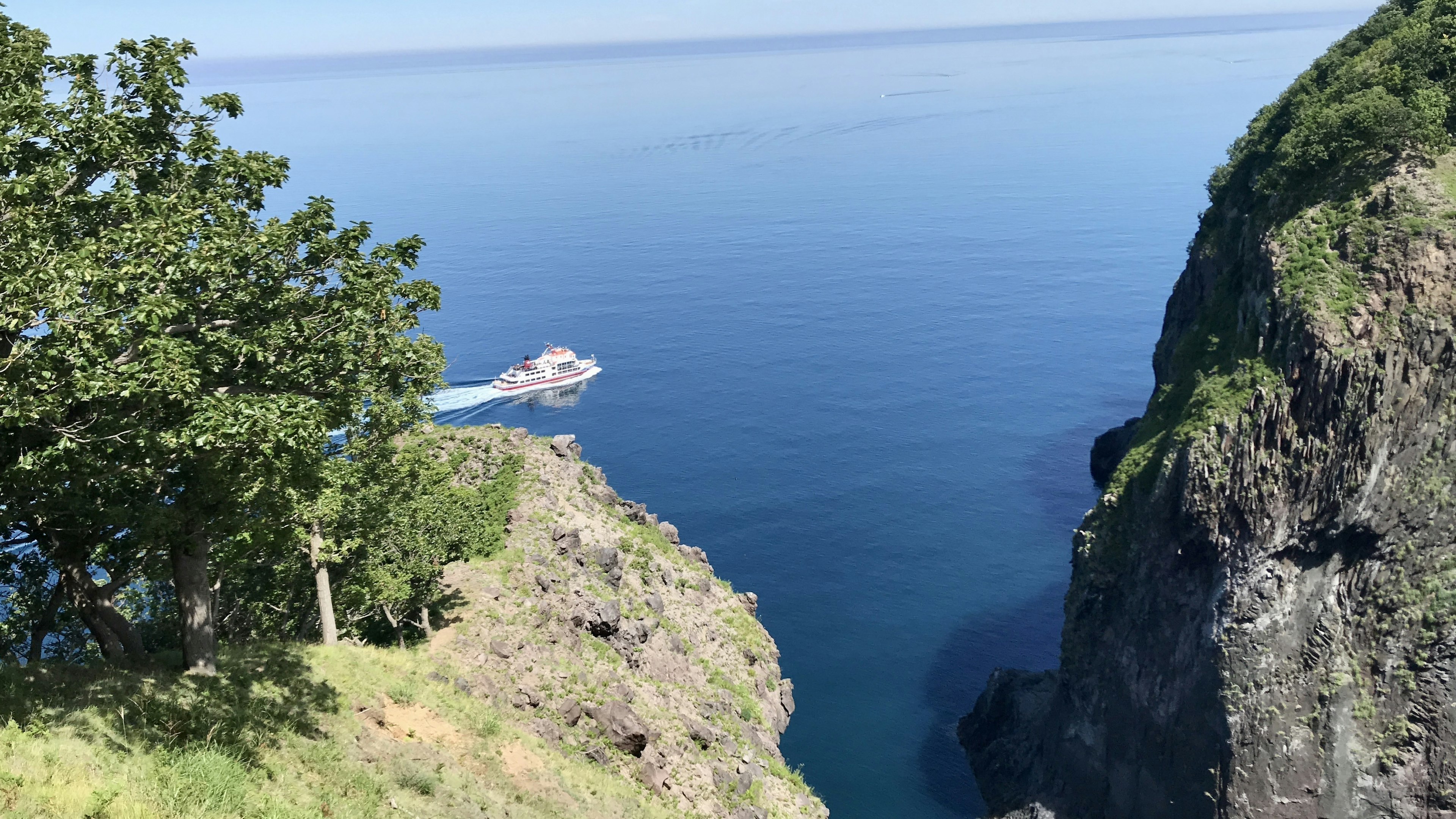 Scenic view of cliffs with green trees overlooking a blue sea and a boat navigating through the water