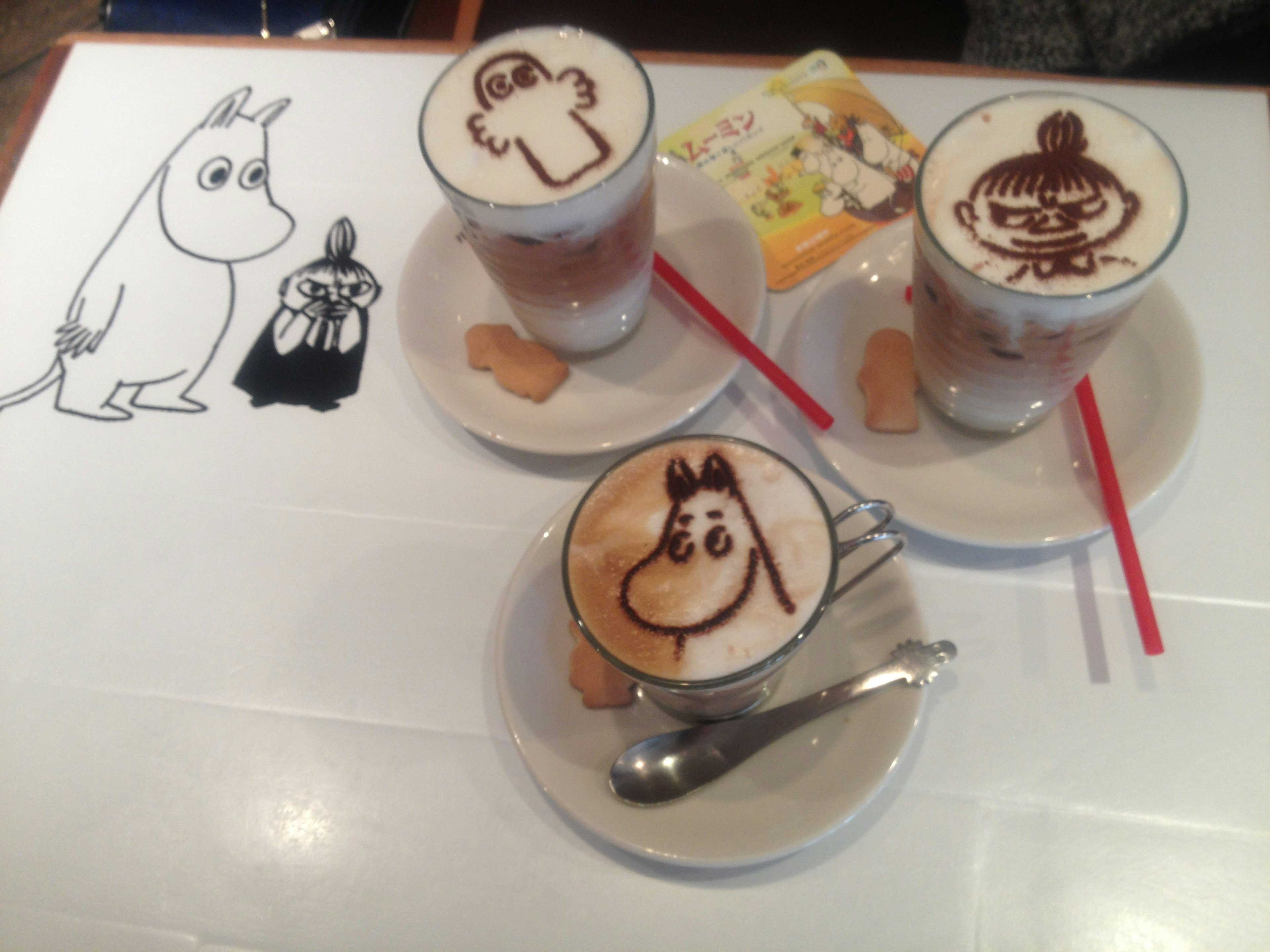 Tiga cangkir dengan seni latte karakter Moomin di atas meja yang memiliki gambar