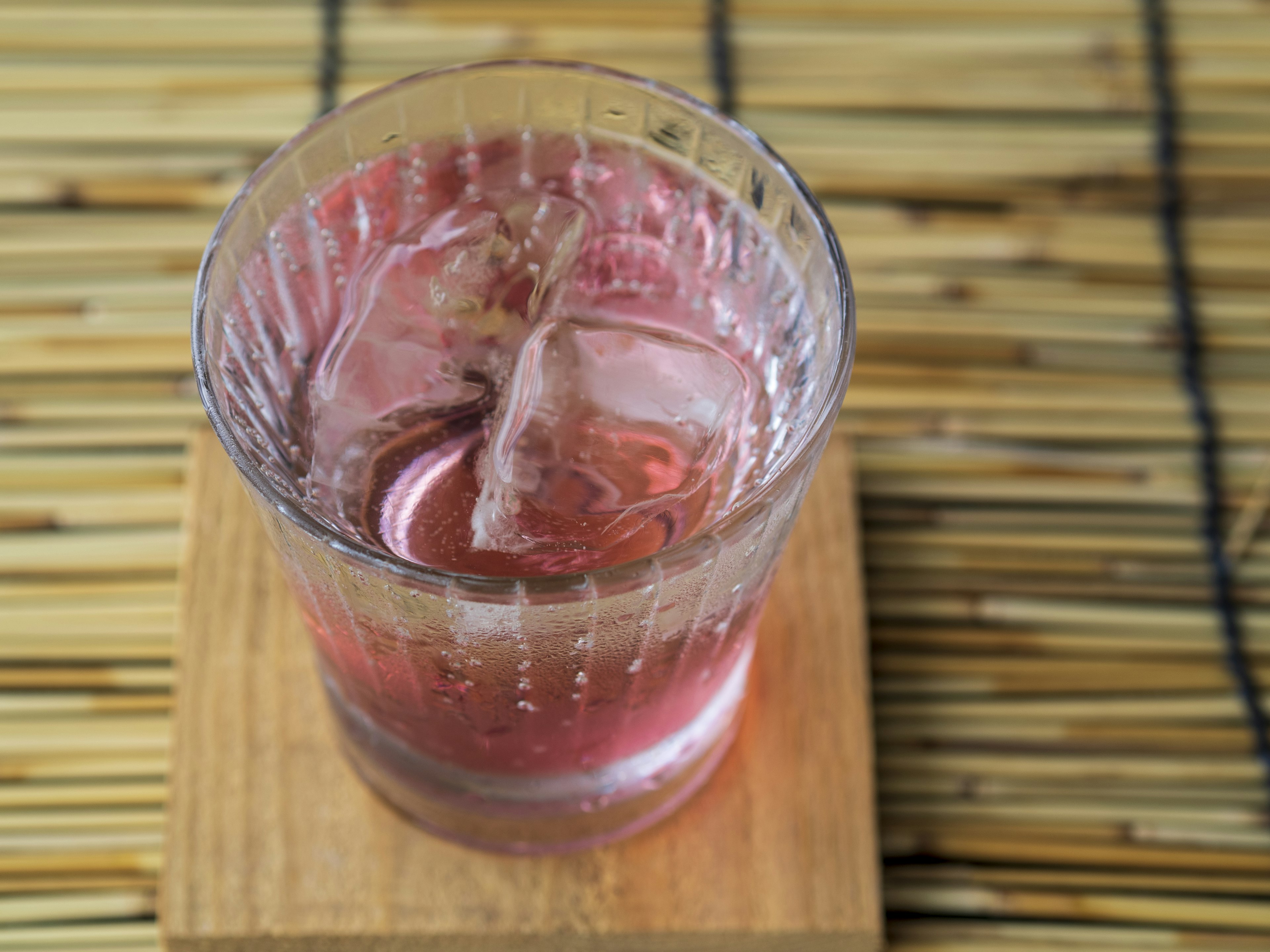 A pink drink with ice in a clear glass on a wooden coaster