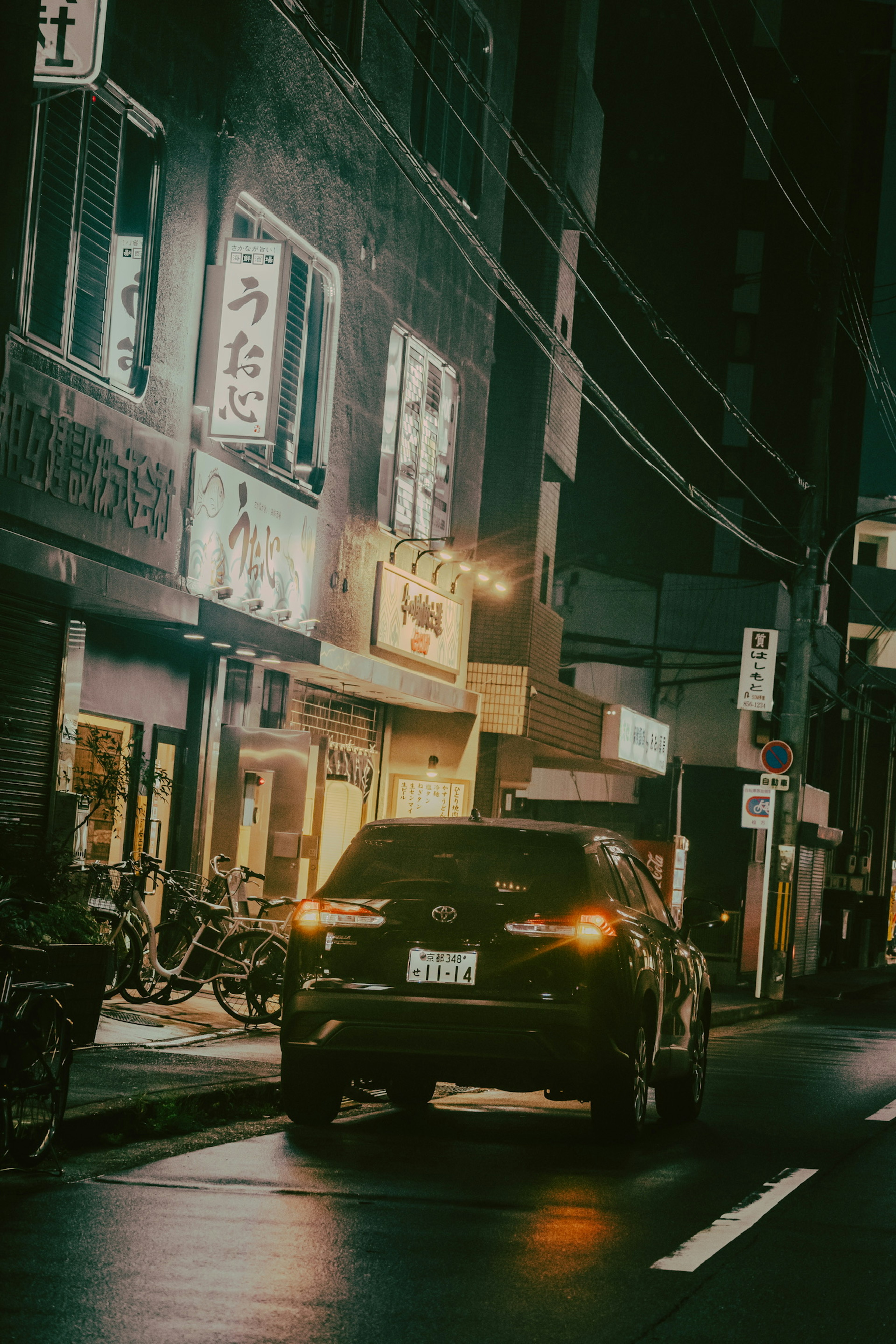 Black car parked on a dimly lit street with neon signs and shops