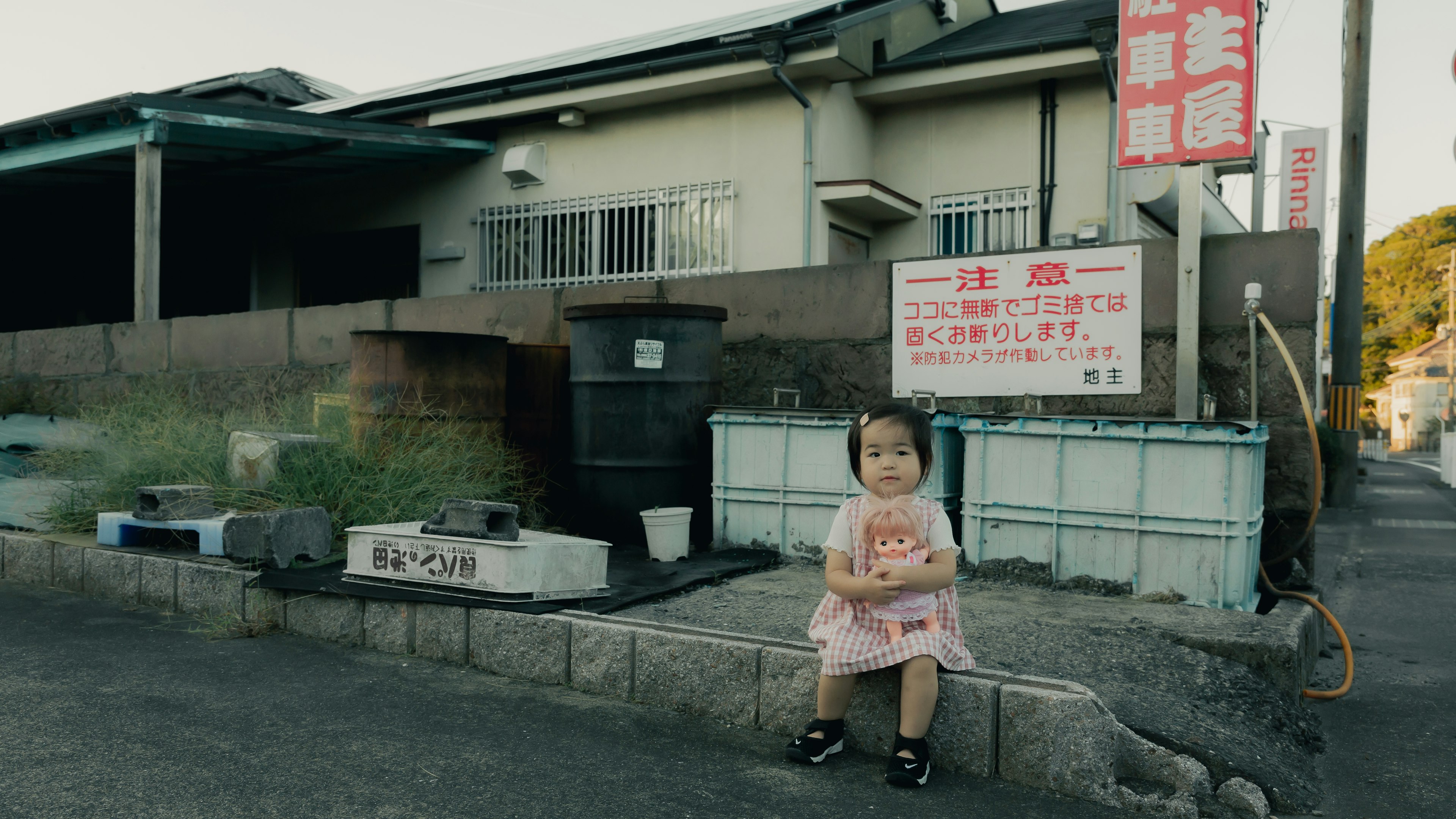 Una niña pequeña sentada al borde de la carretera frente a una casa japonesa con un cartel y tanques de agua