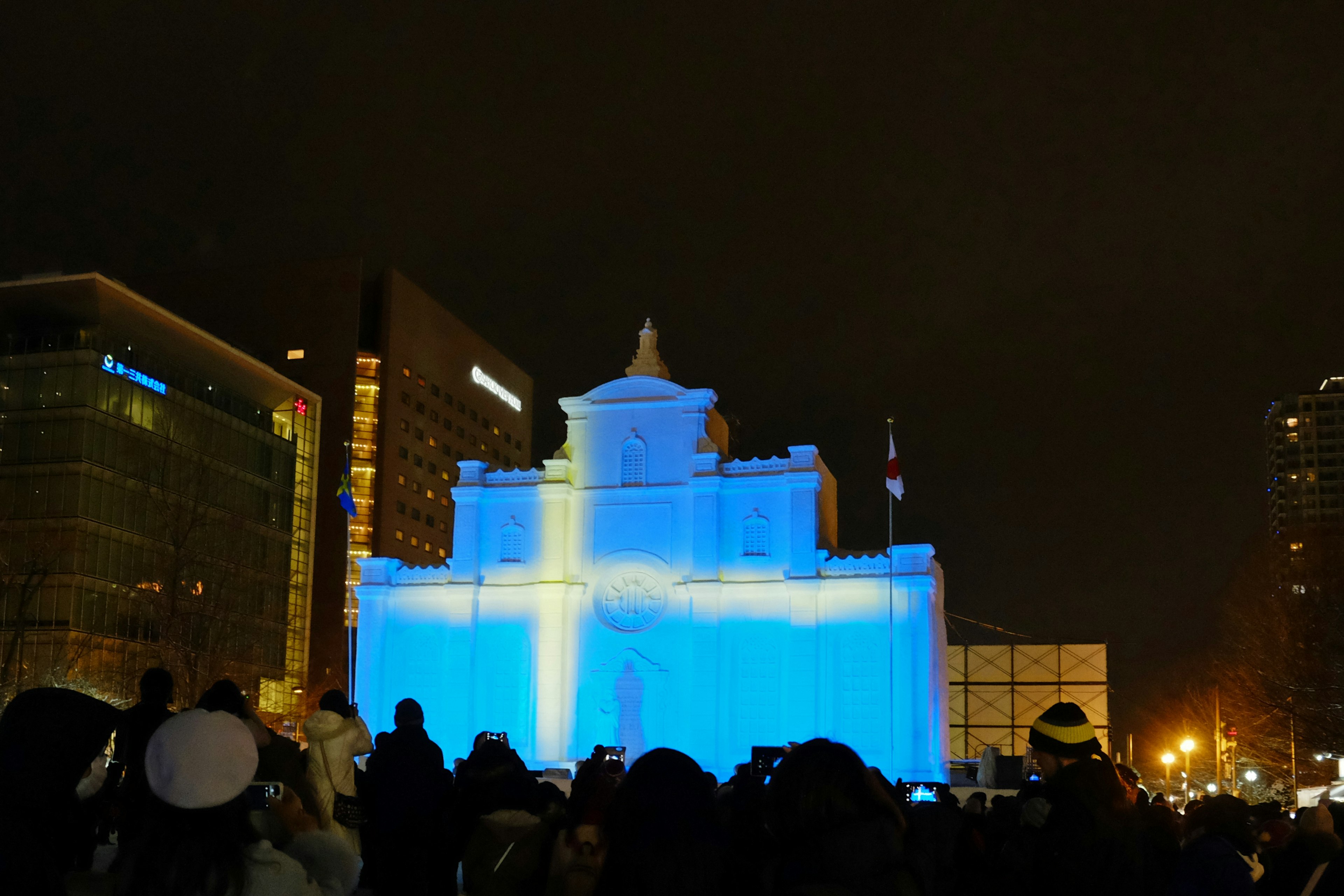 Building illuminated with a blue cross projection at night with a crowd