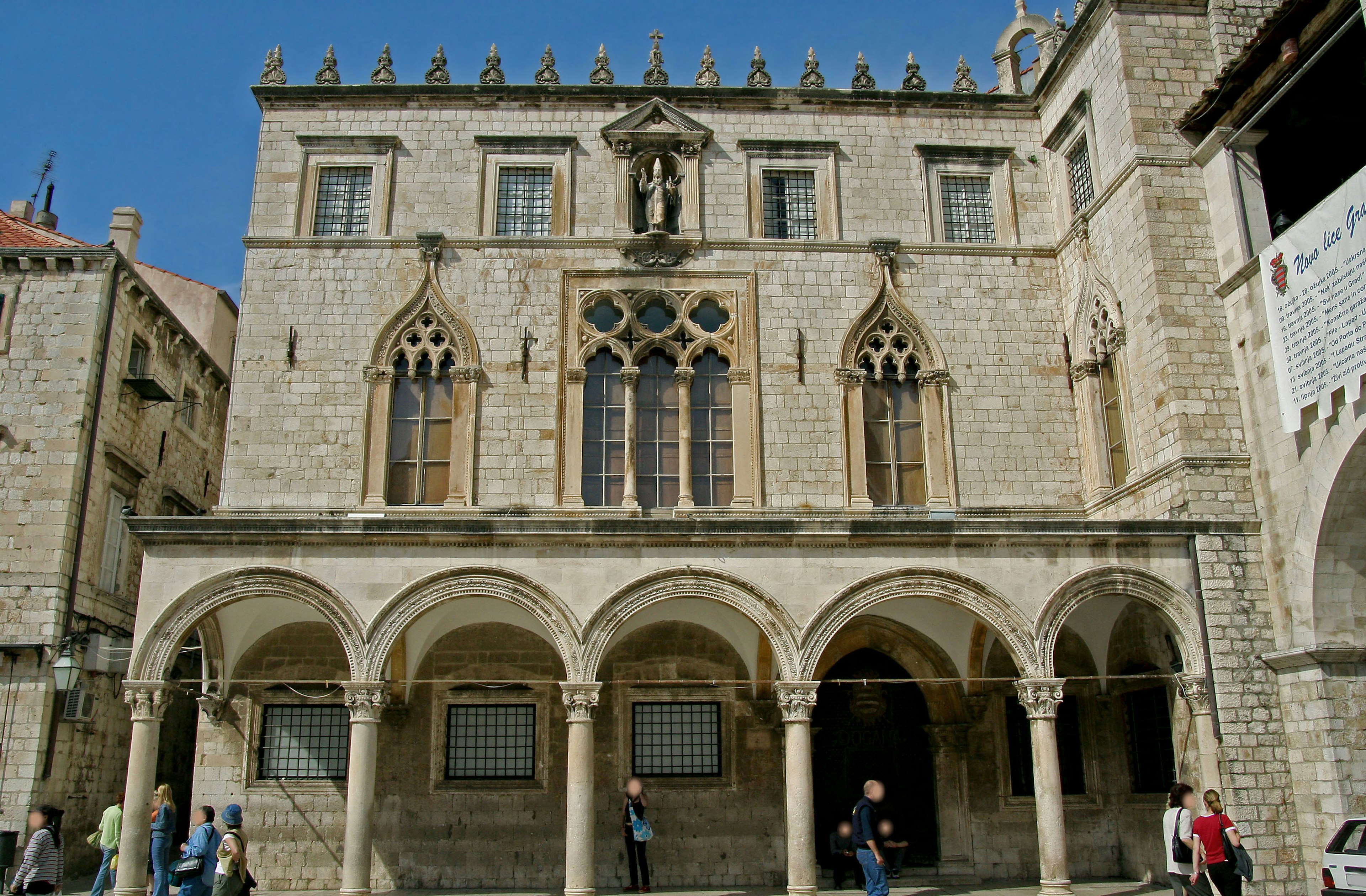 Edificio histórico en Dubrovnik con ventanas góticas y columnas arqueadas bajo un cielo azul con turistas