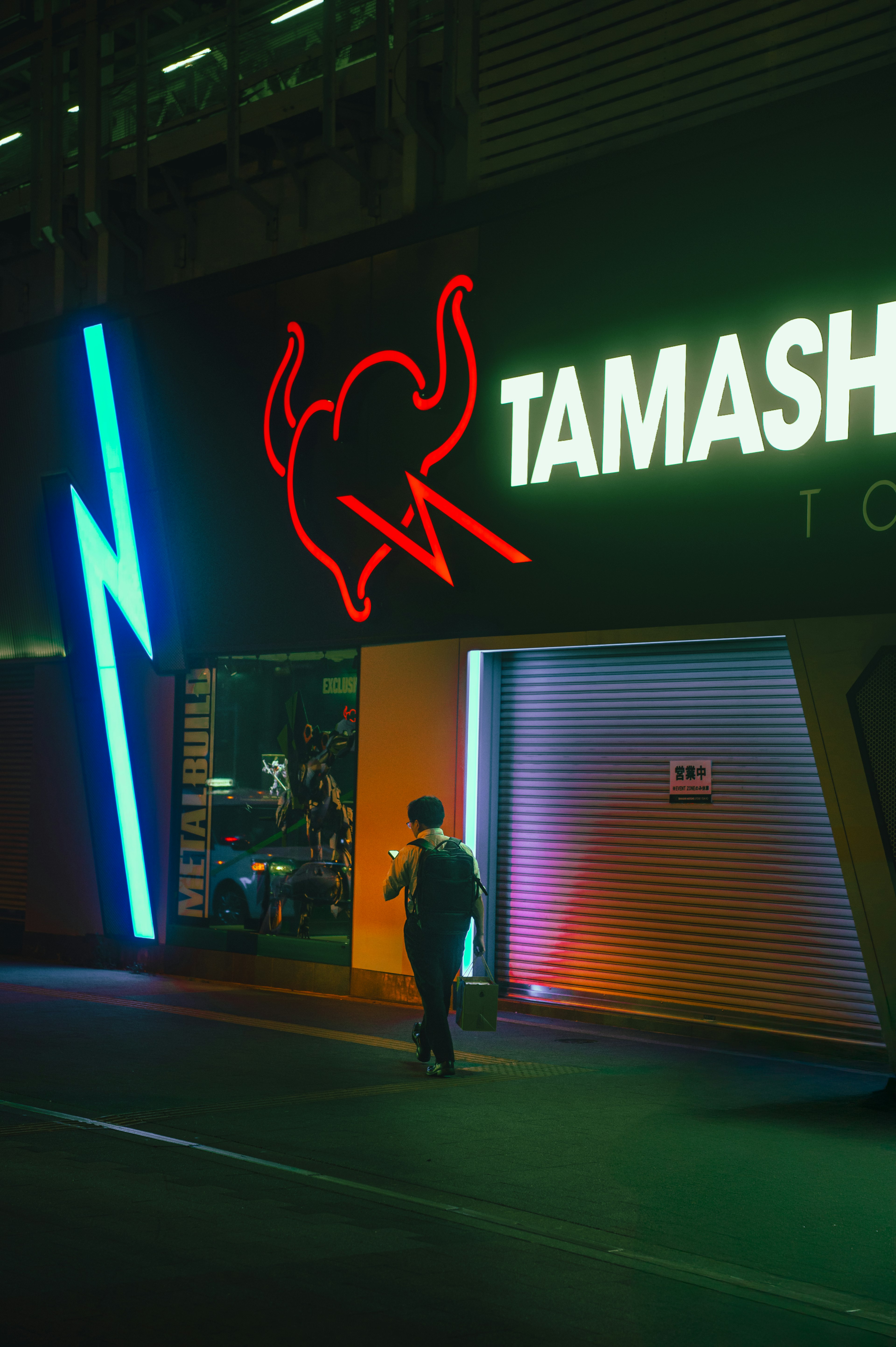 Person walking in front of illuminated Tamashii Tokyo store with neon signs