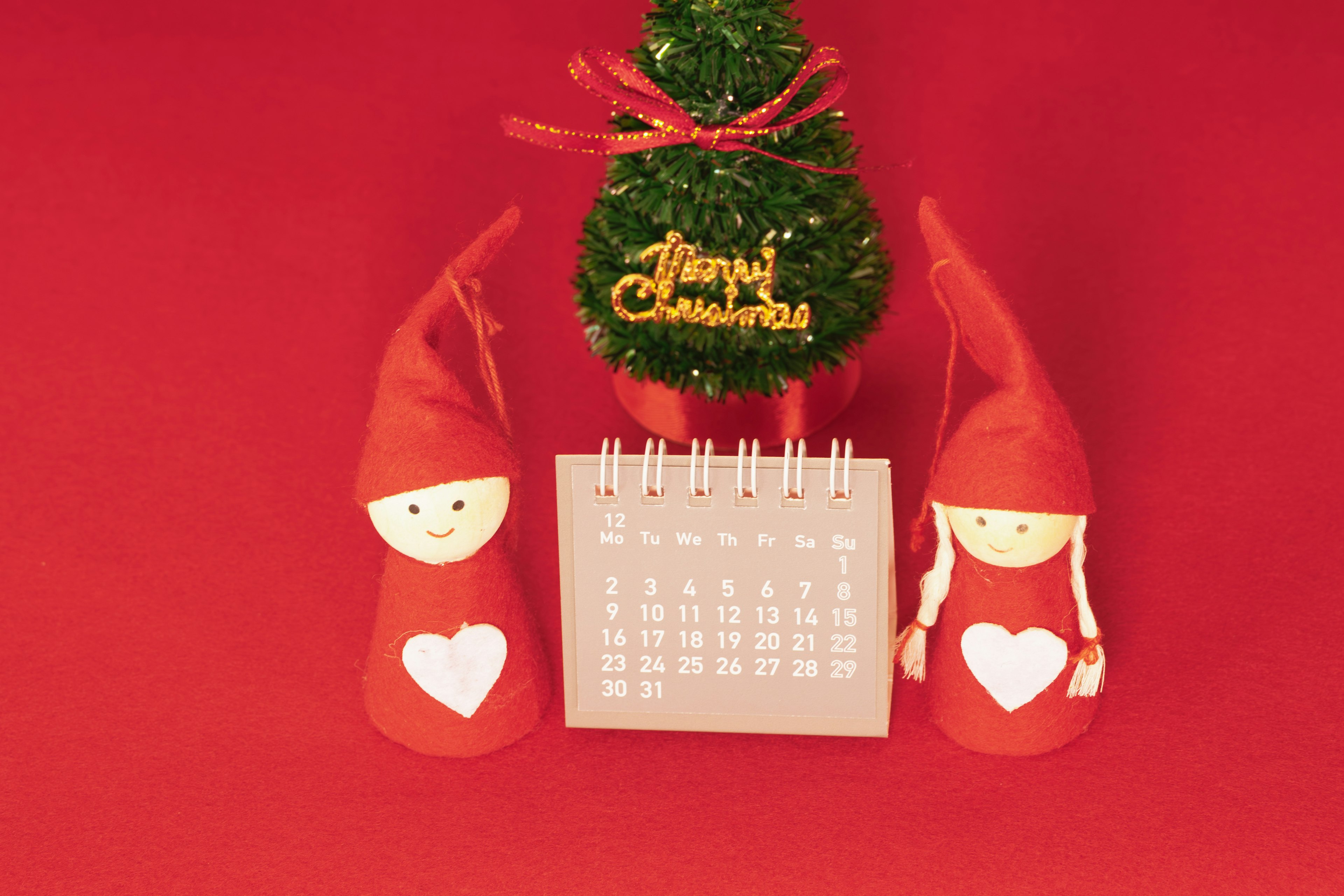 Two red-hatted dolls with hearts beside a small Christmas tree and calendar