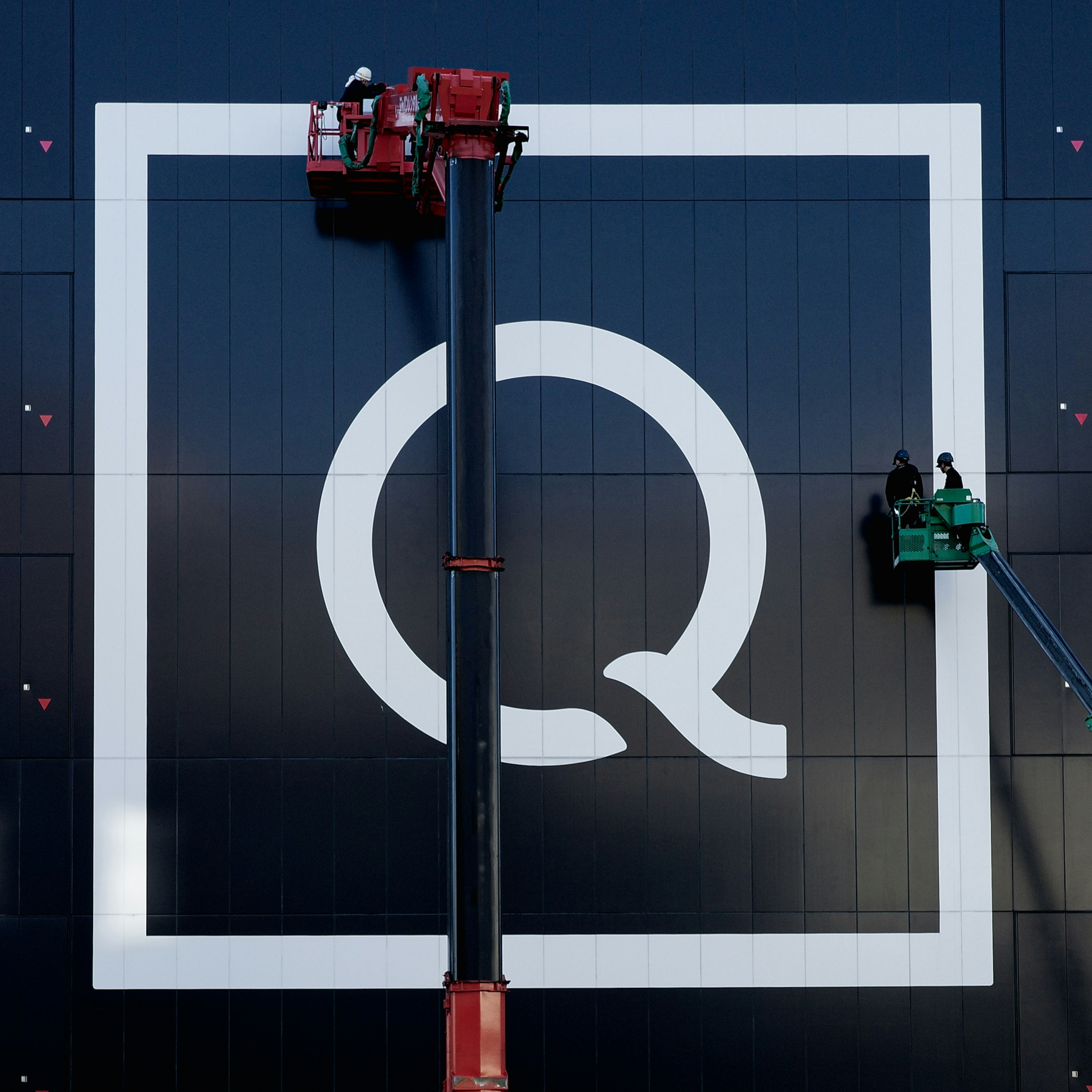 Large white Q logo on black wall with workers using cherry pickers