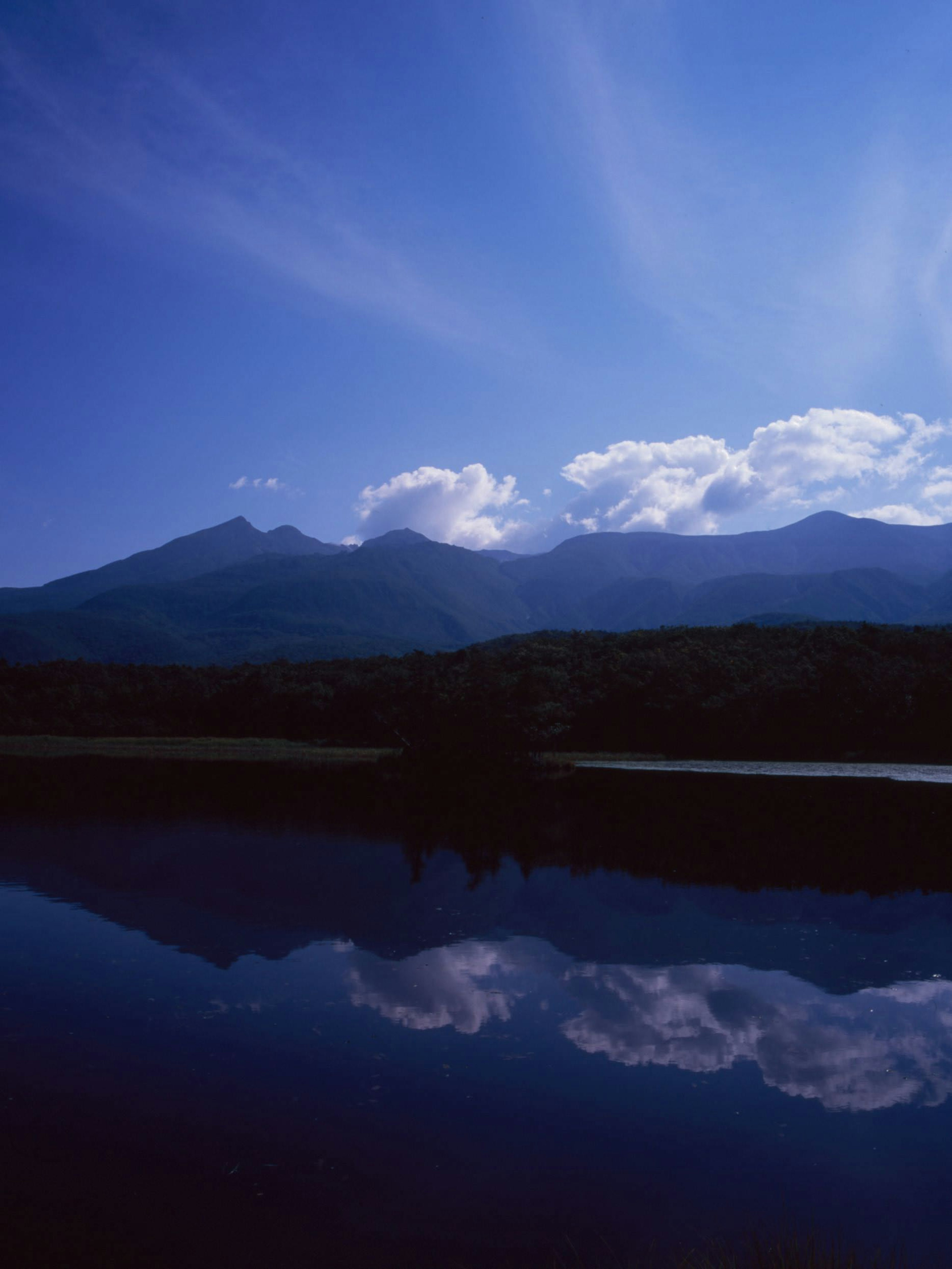 Danau tenang yang memantulkan langit biru dan awan dengan pegunungan di latar belakang yang menunjukkan keindahan alam