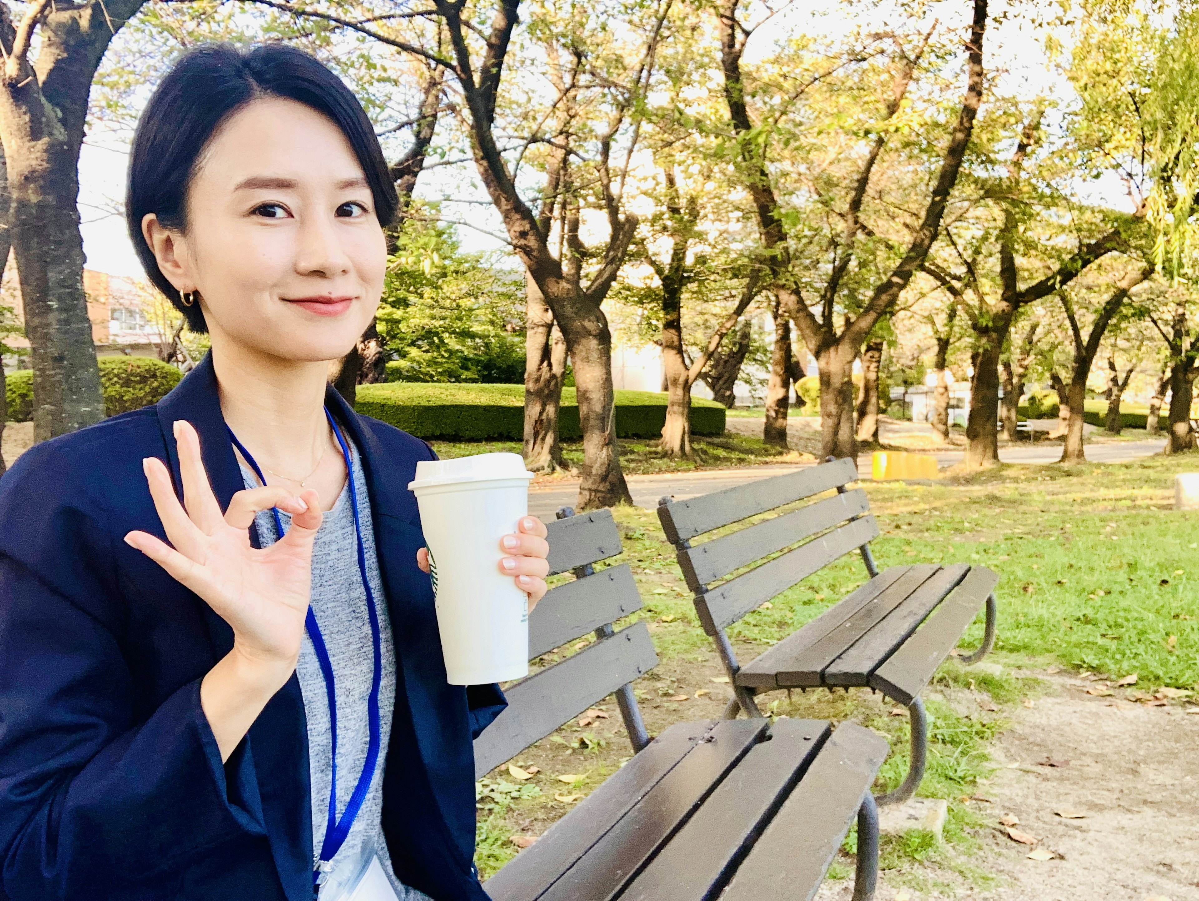 A woman sitting on a park bench holding coffee and giving an OK sign