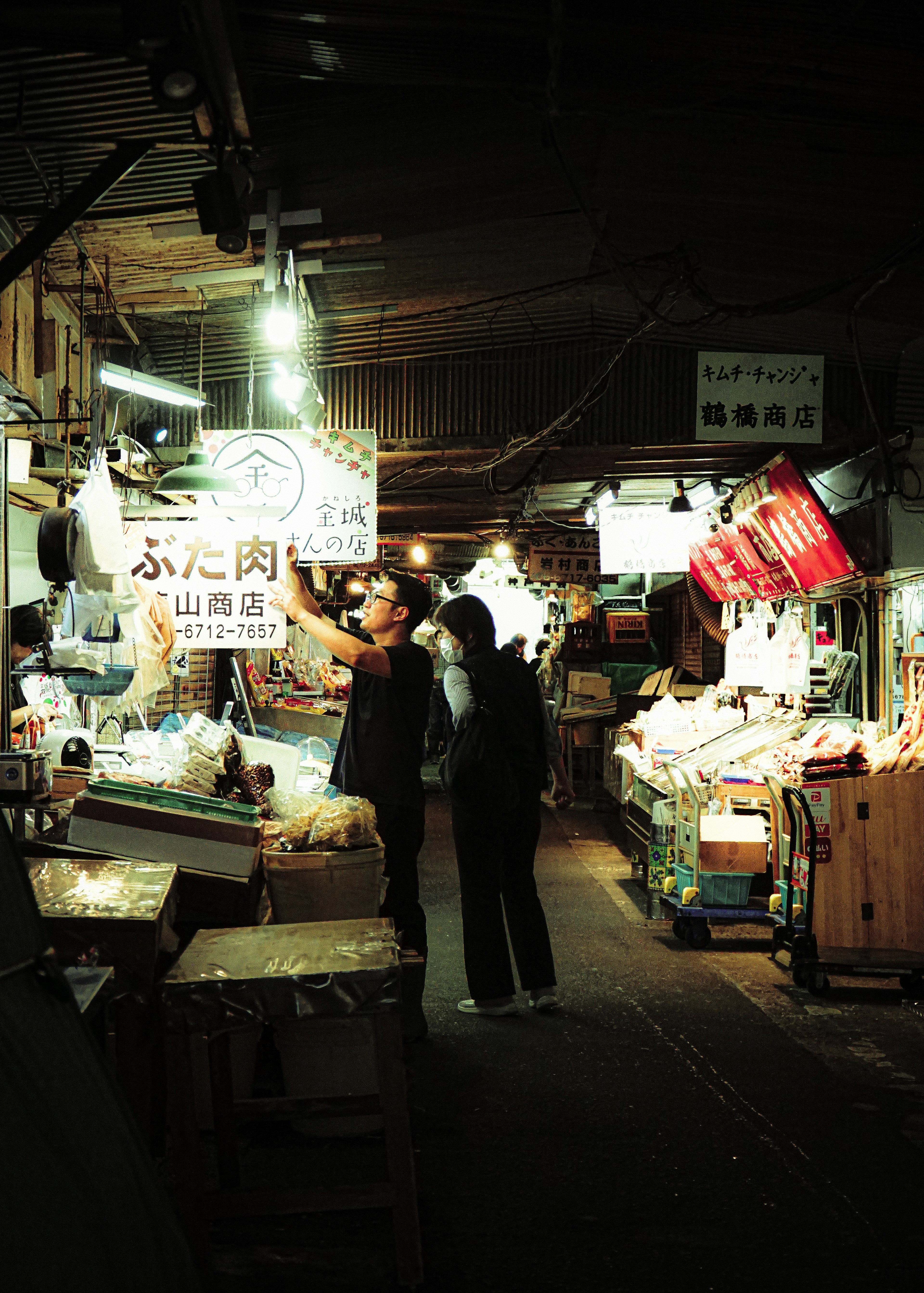 People selecting goods in a dimly lit market aisle with bright signs