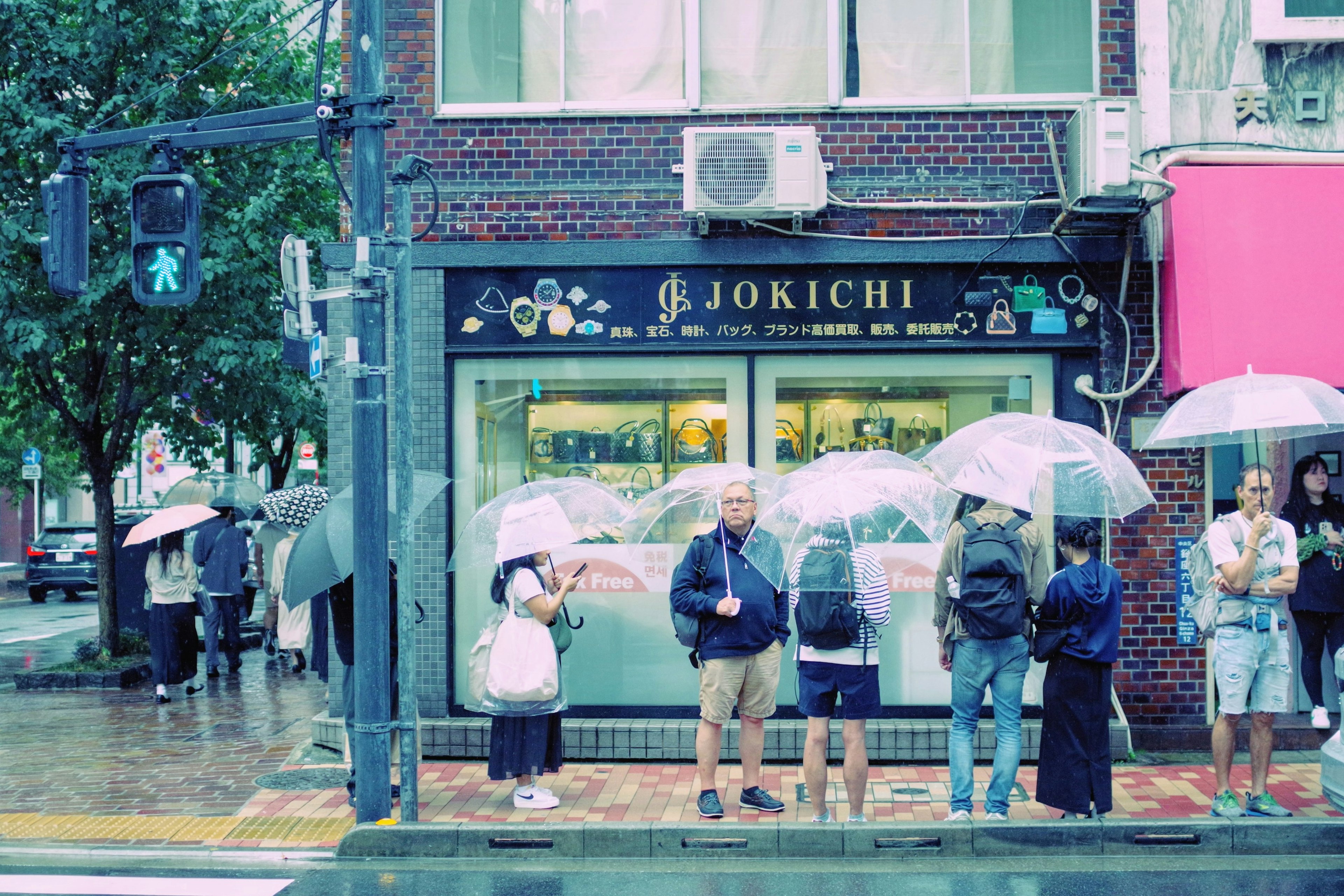 雨の日に傘を持って待っている人々とJOKICHIの看板がある風景