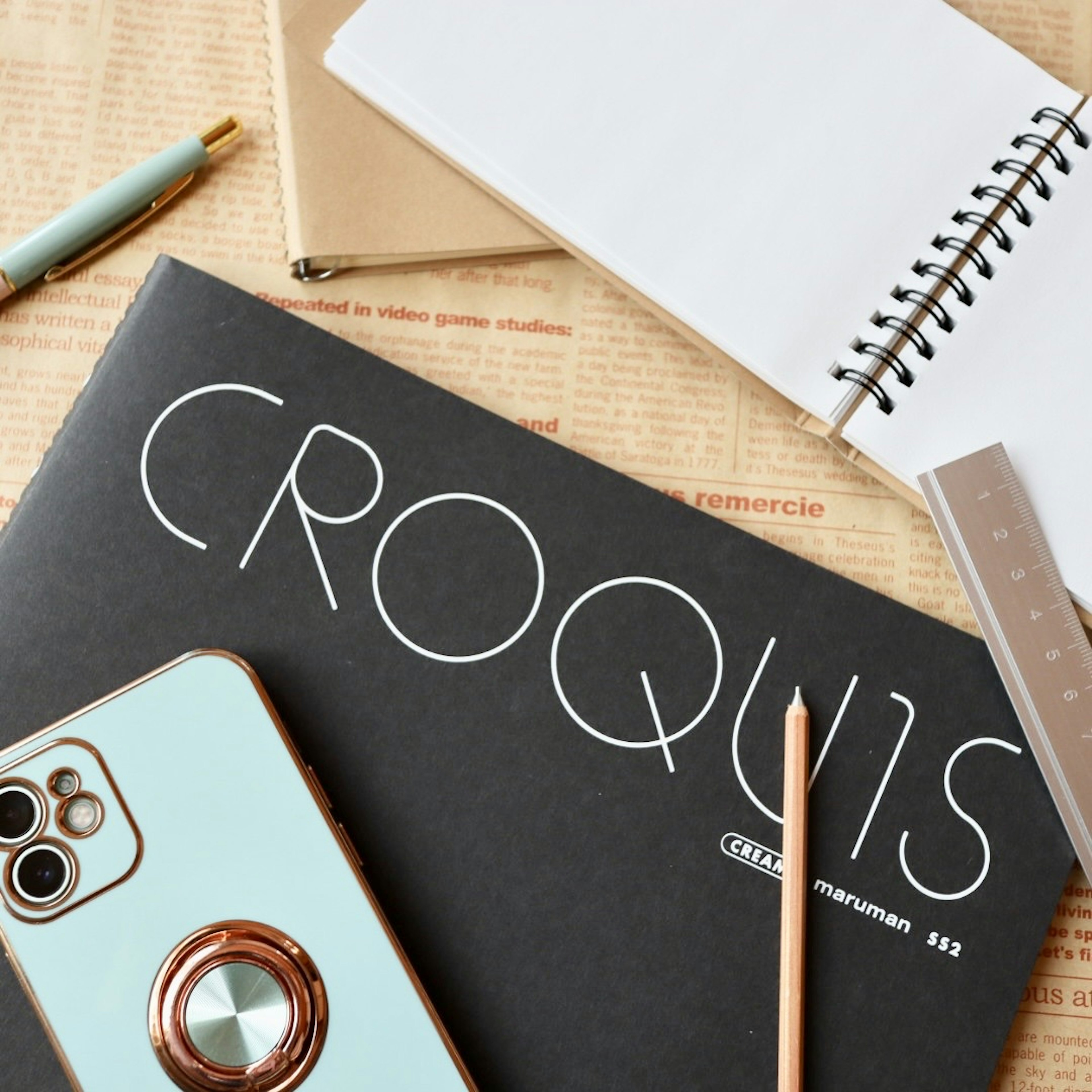 A black sketchbook with the word CROQUIS and various pens on a desk with a smartphone and a notebook