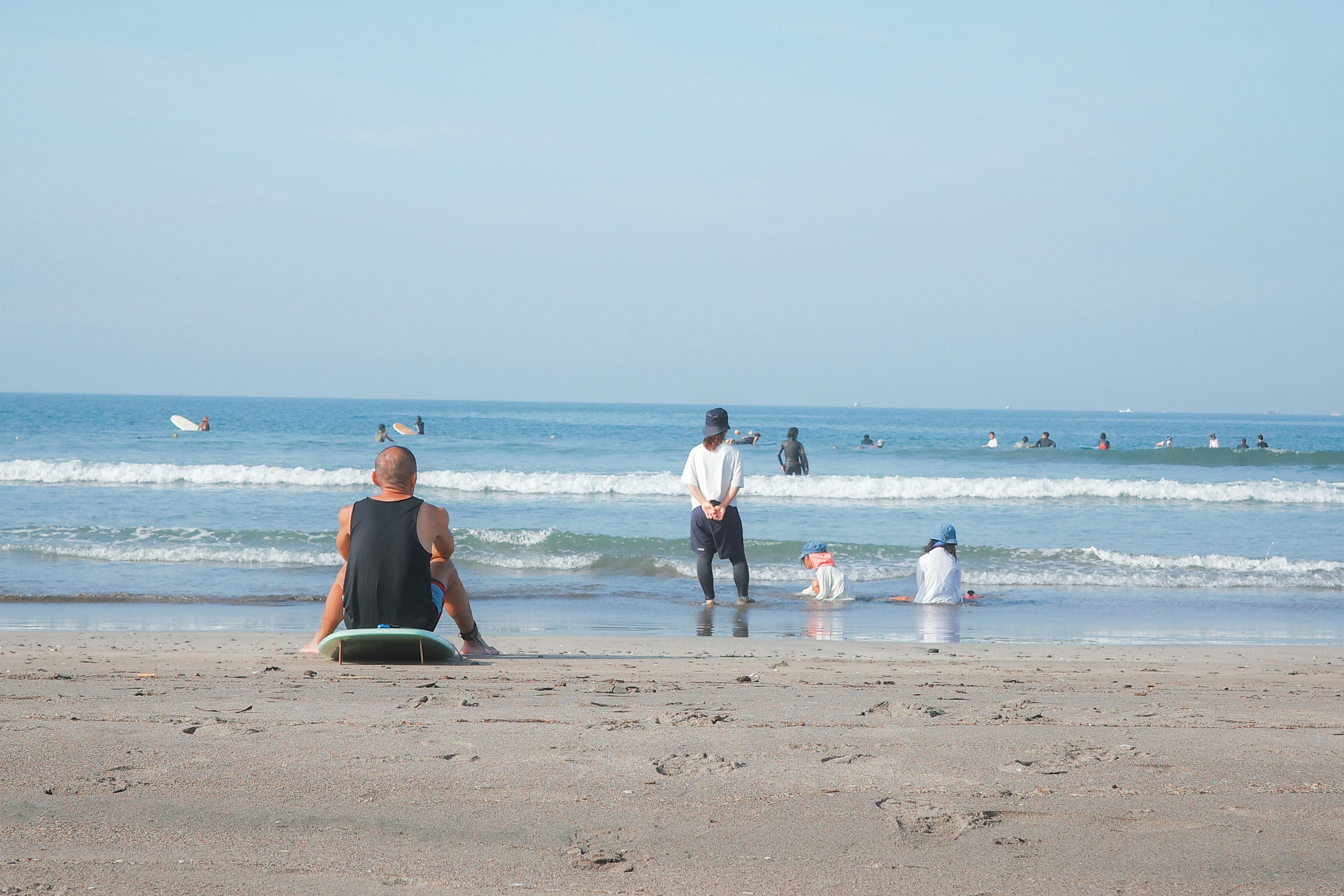 A beach scene with a person sitting on the sand waiting to surf and children playing in the water