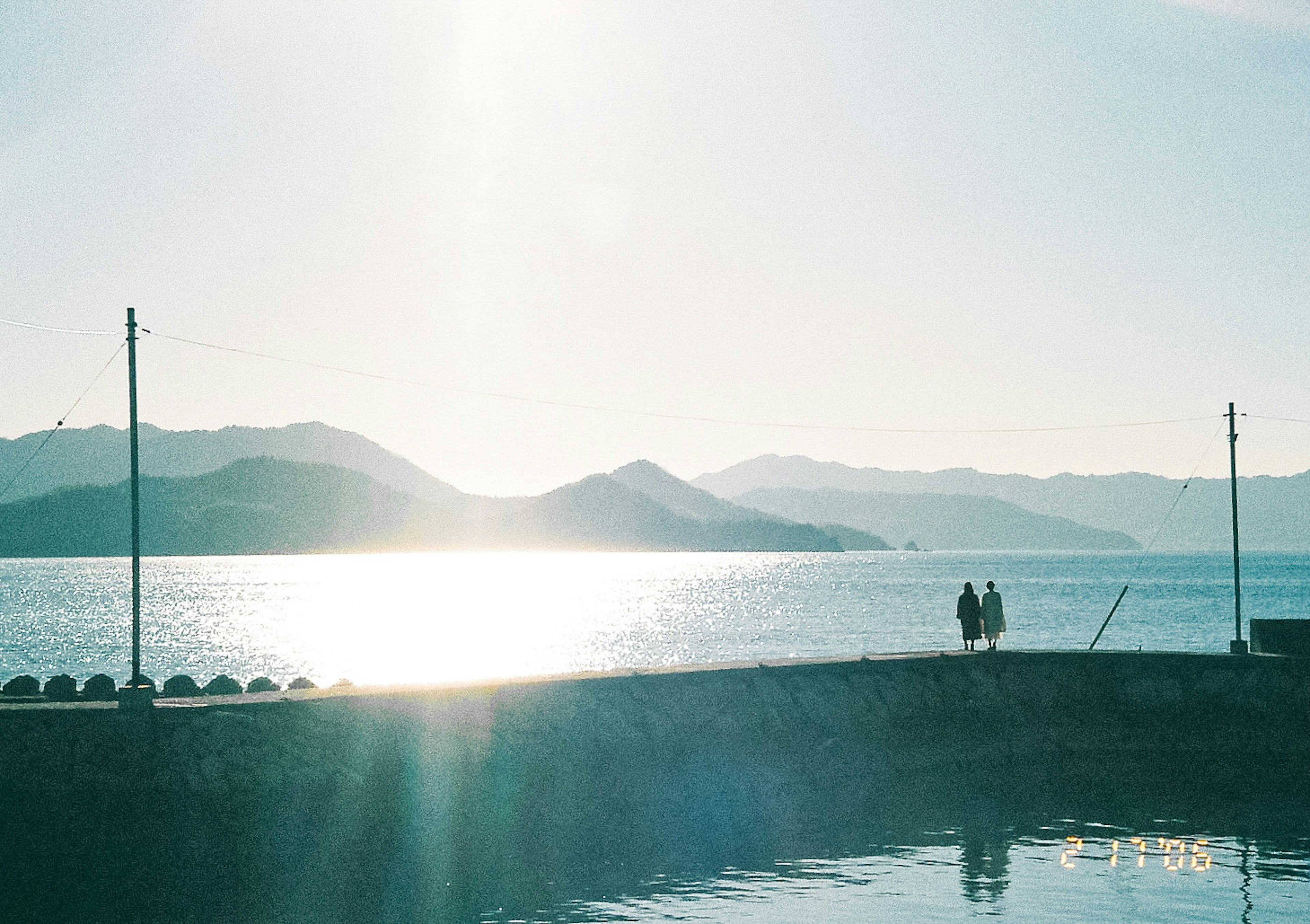 穏やかな海と山々を背景にした夕焼けの景色で、二人の人が海岸に立っている