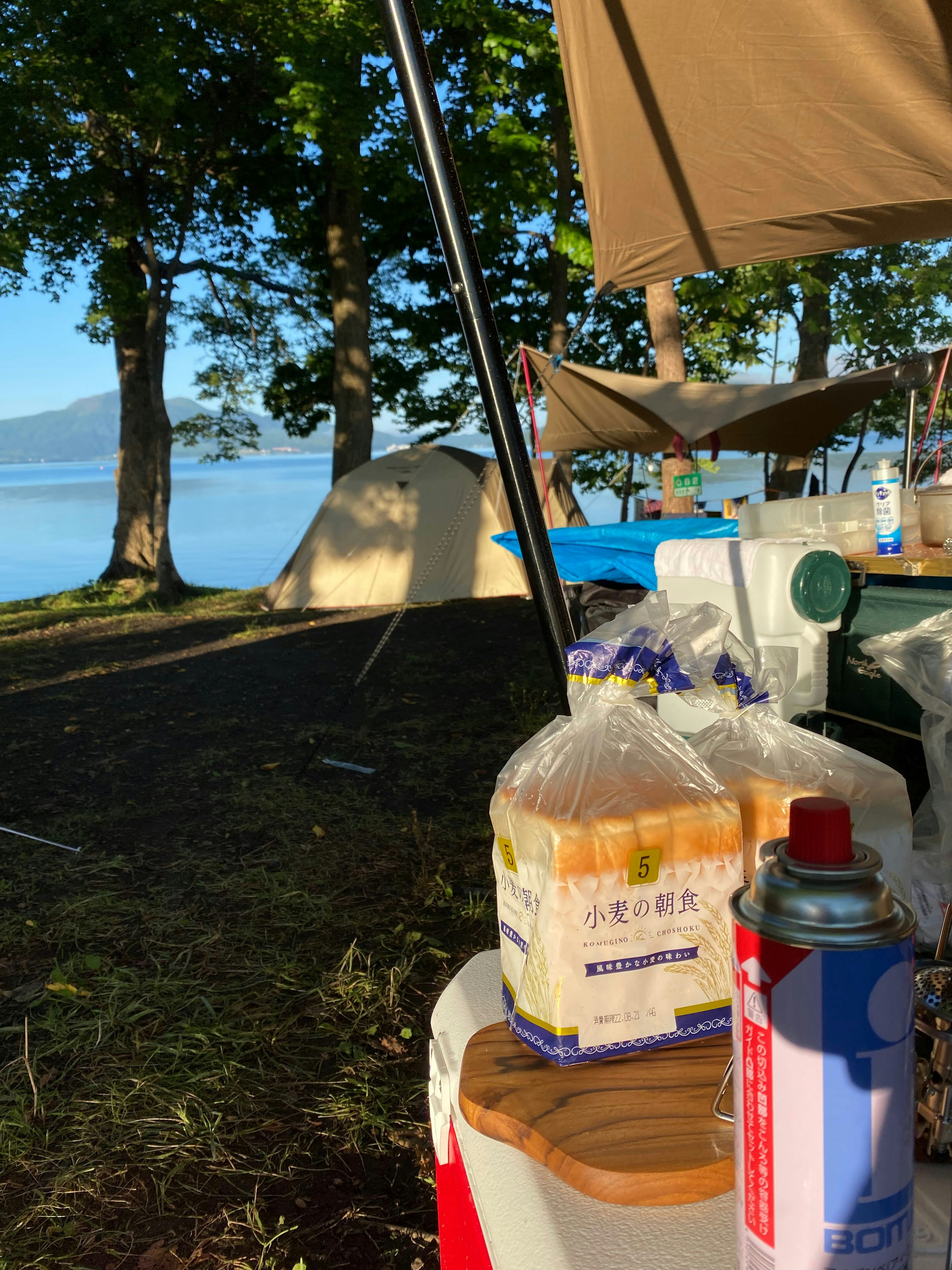 Vista de suministros de camping que incluyen pan y spray de cocina en una mesa con un lago y árboles al fondo