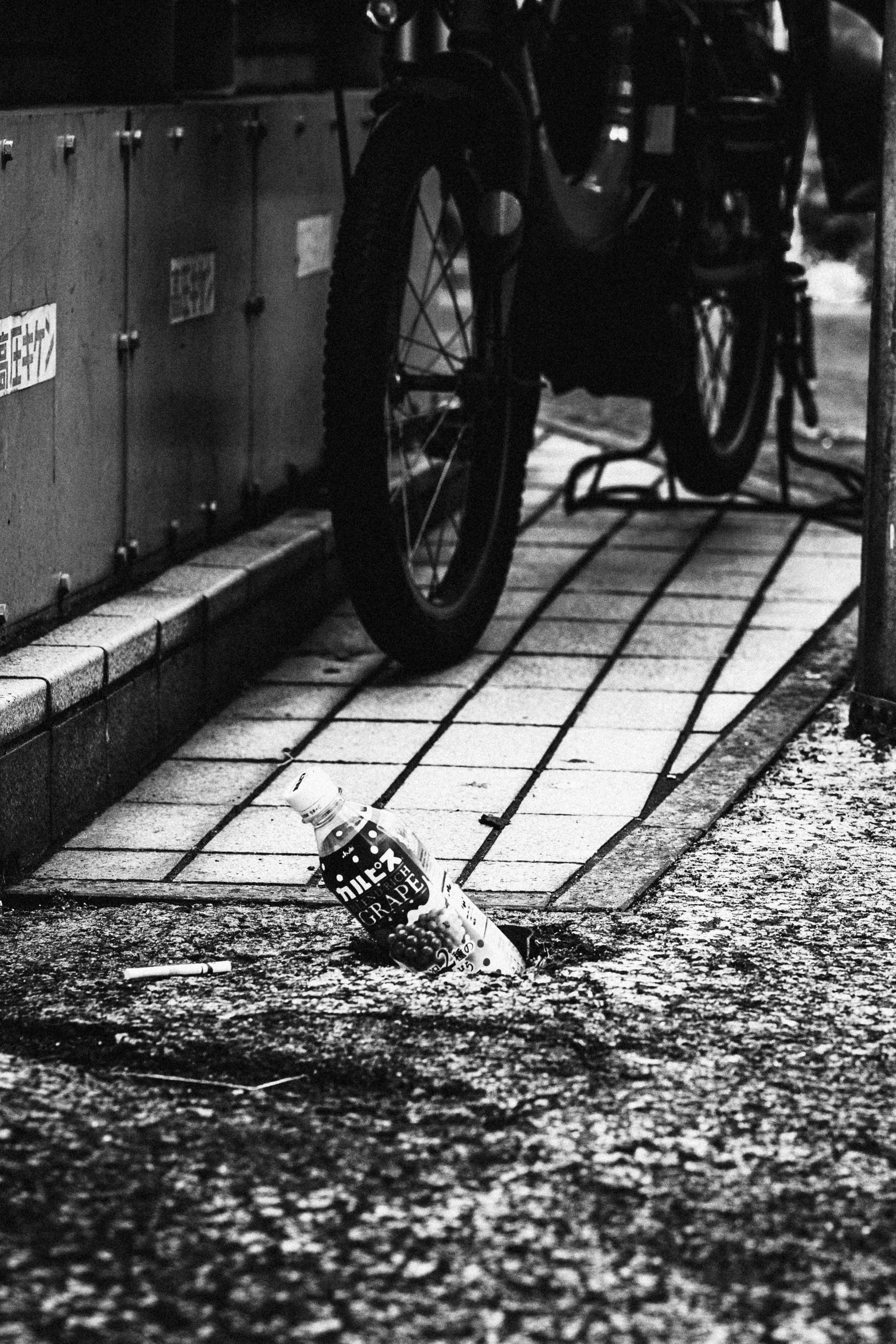 Black and white photo of a discarded bottle on the ground with a bicycle wheel in the background