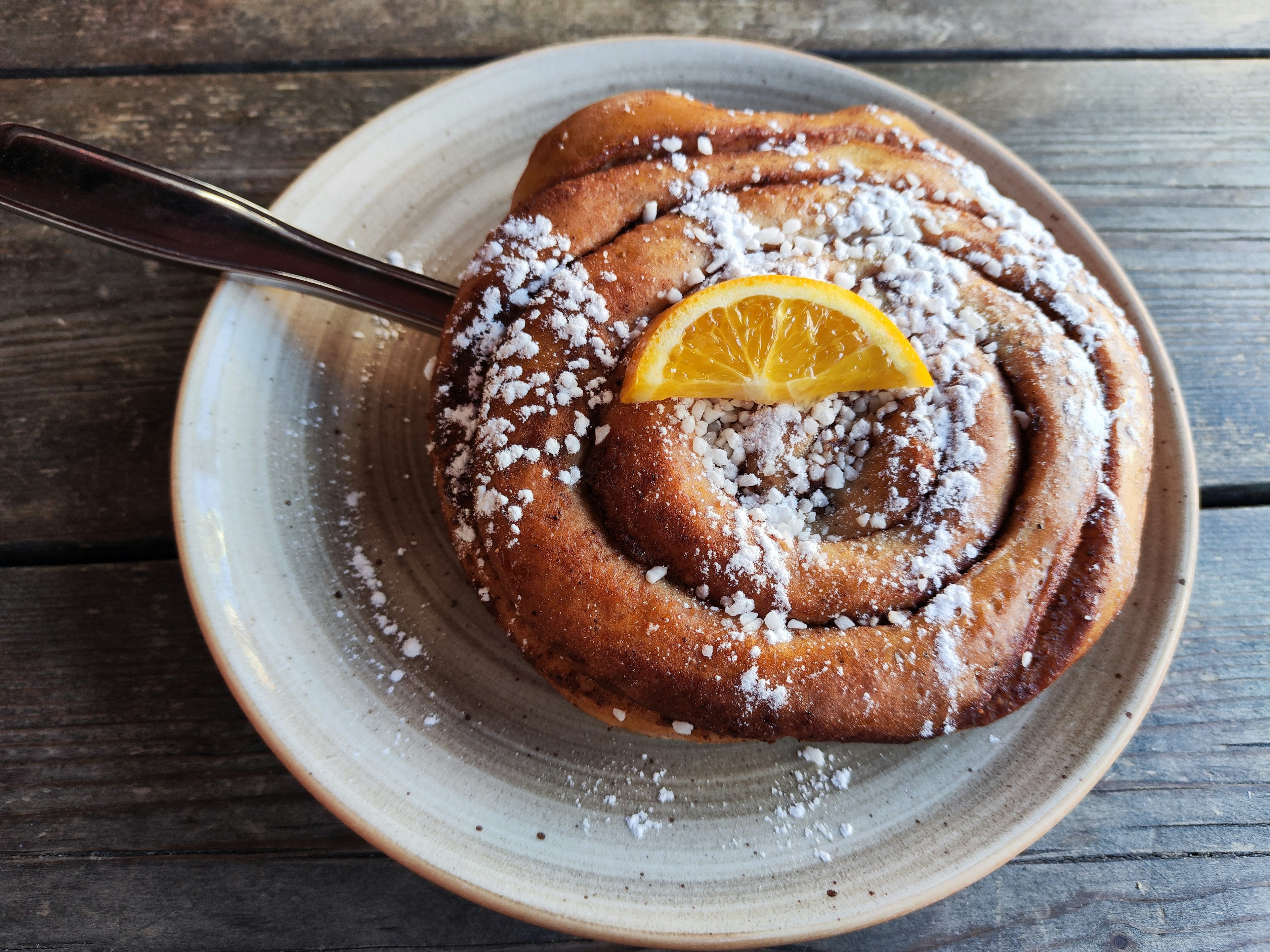 Cinnamon roll topped with powdered sugar and a slice of orange on a rustic plate