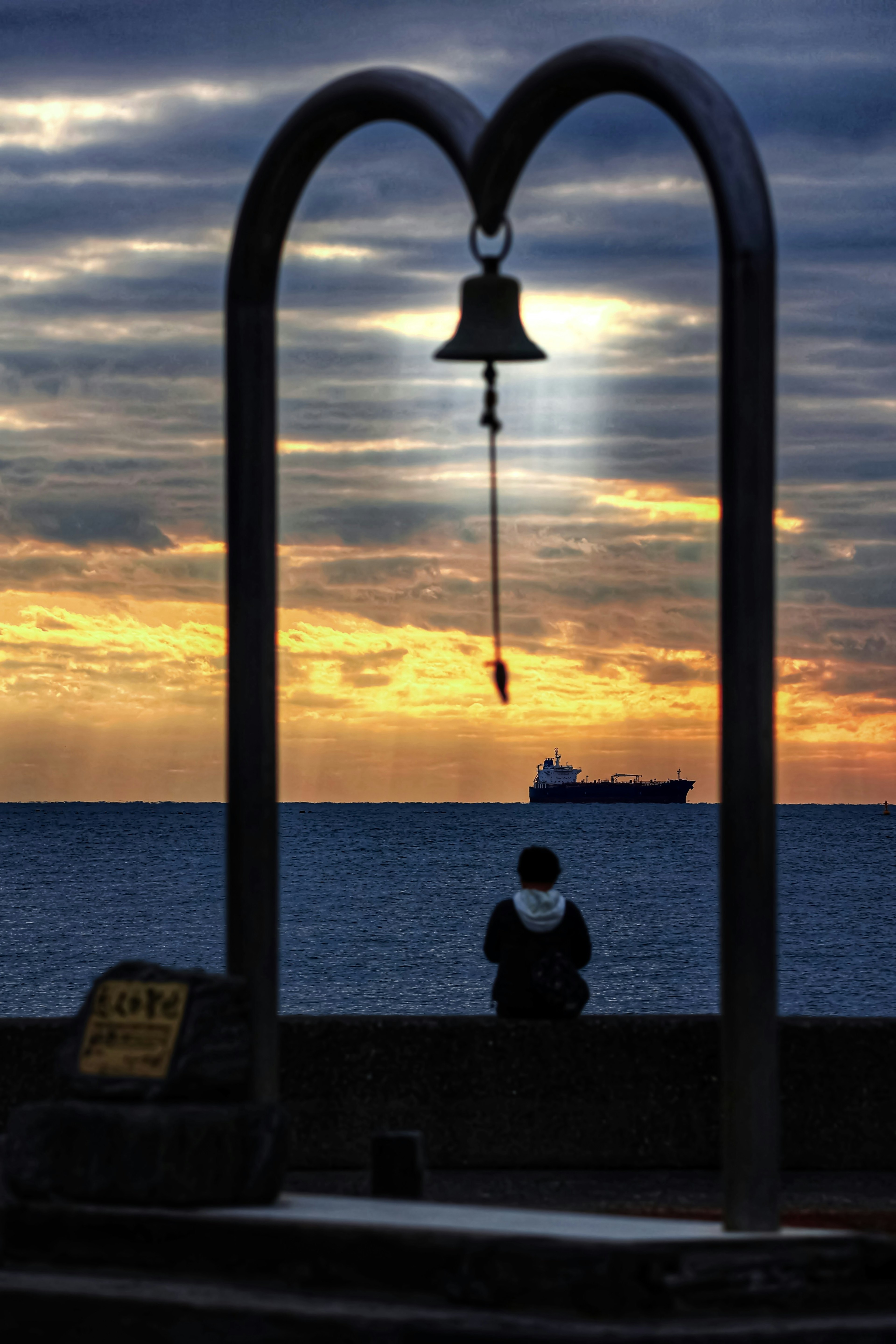 Silhouette einer Person, die am Meer sitzt, umrahmt von einem Glockenbogen bei Sonnenuntergang