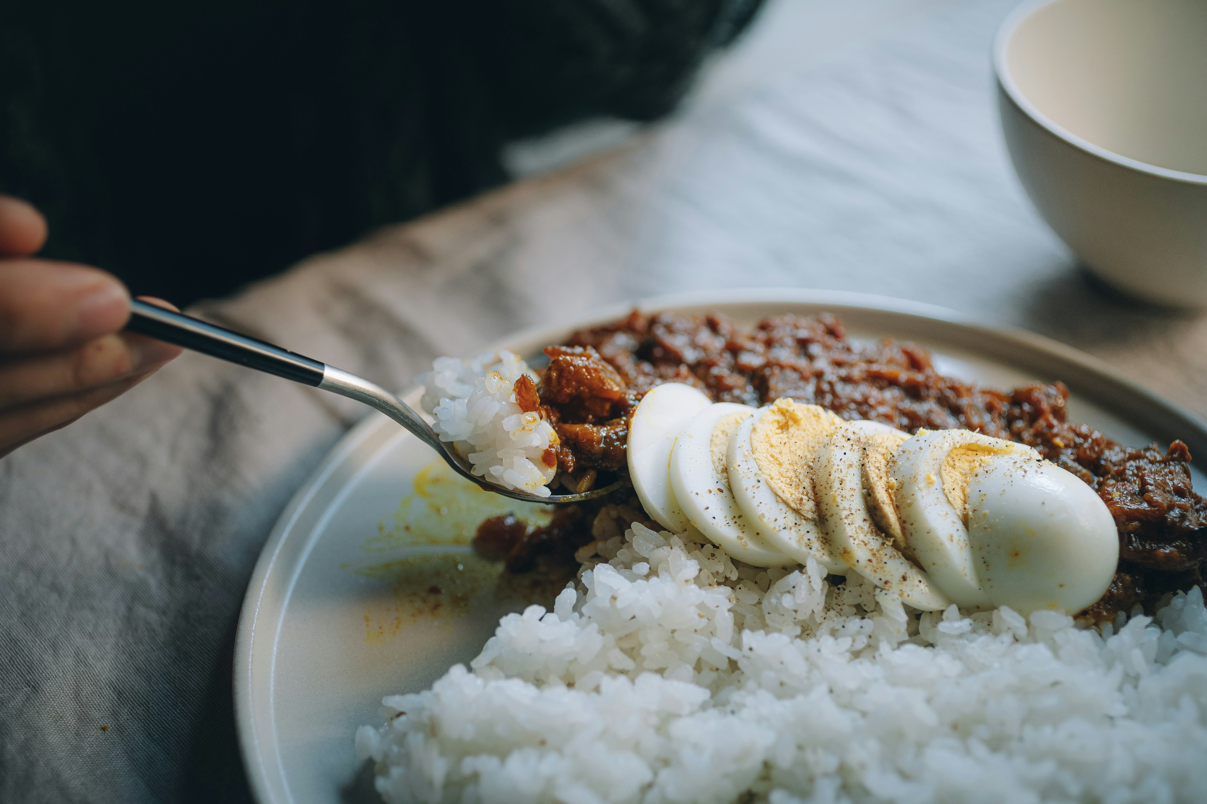 Close-up of a hand using a fork to serve rice topped with sliced eggs and meat