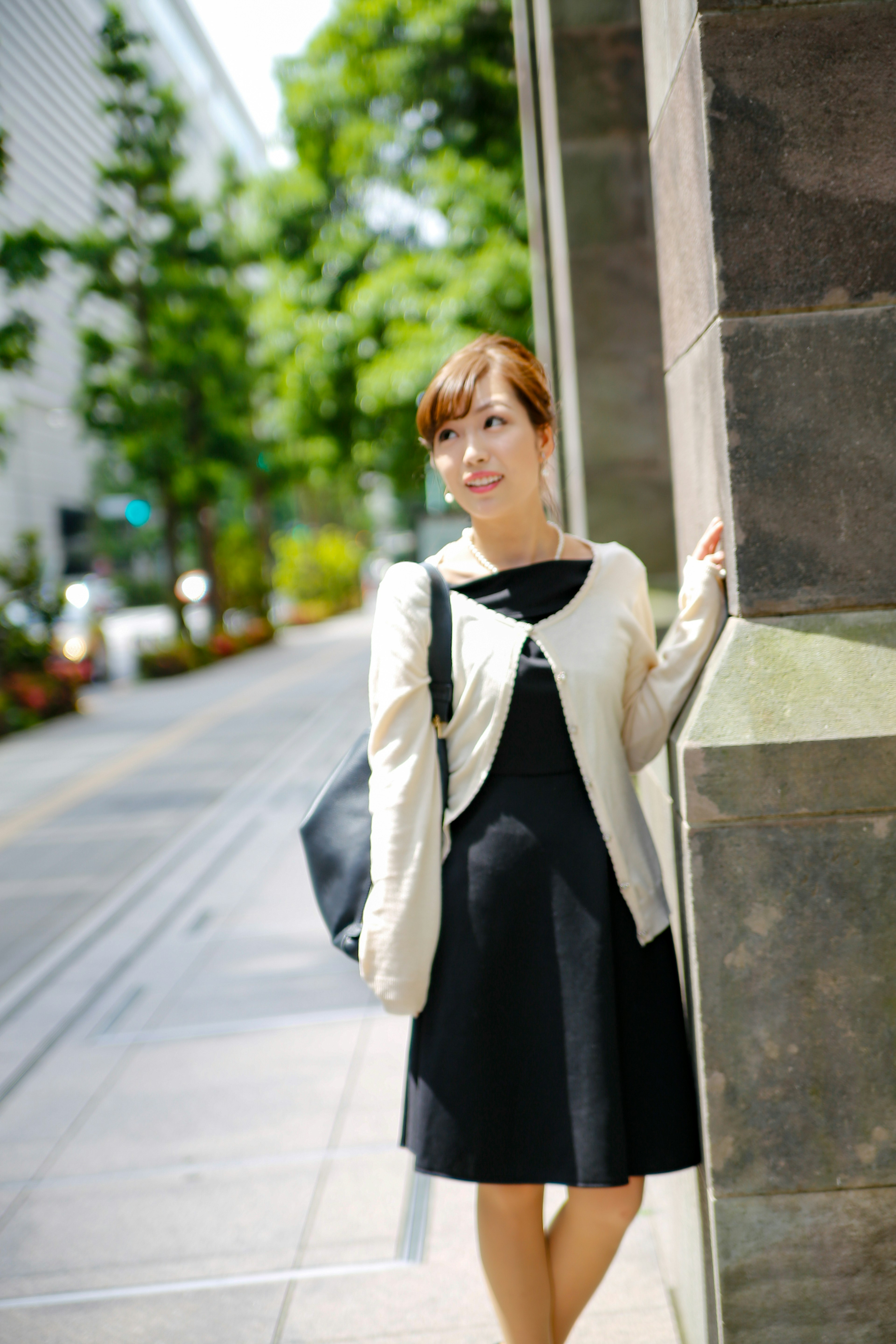 A woman in a black dress and white cardigan stands at a street corner