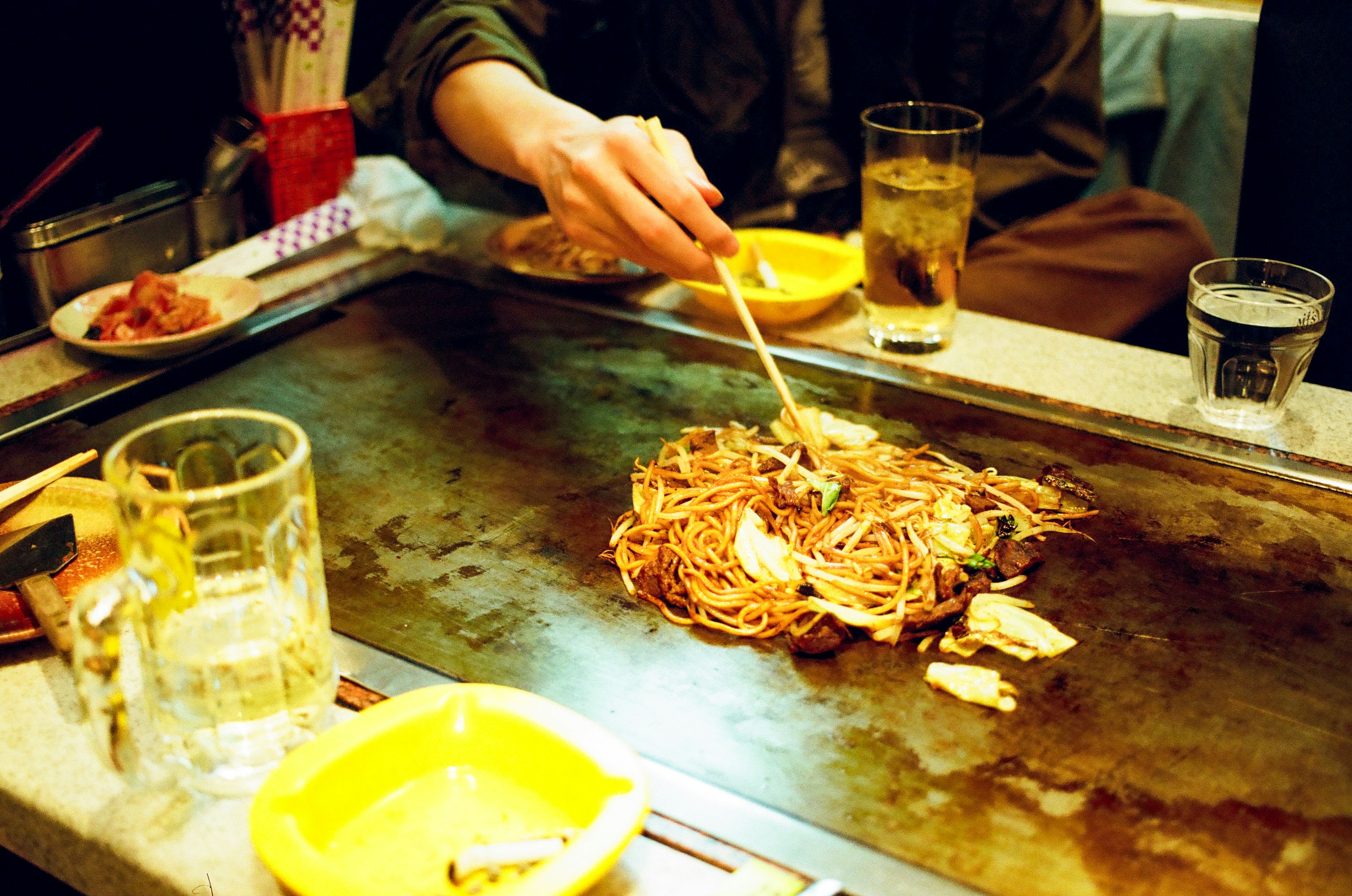 Une main prenant des yakisoba d'une grill teppanyaki avec des boissons sur la table