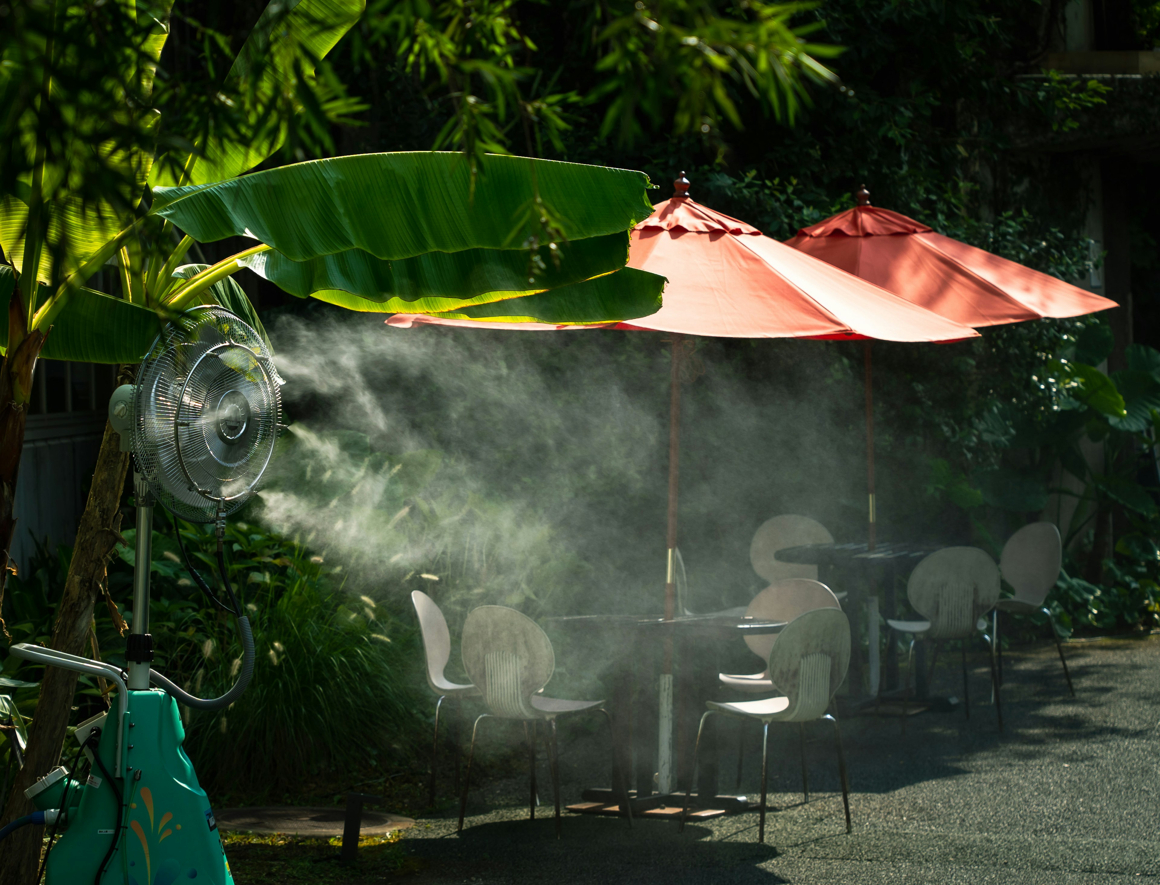 Kipas kabut menyemprotkan uap air di dekat meja kafe dengan daun hijau dan payung merah