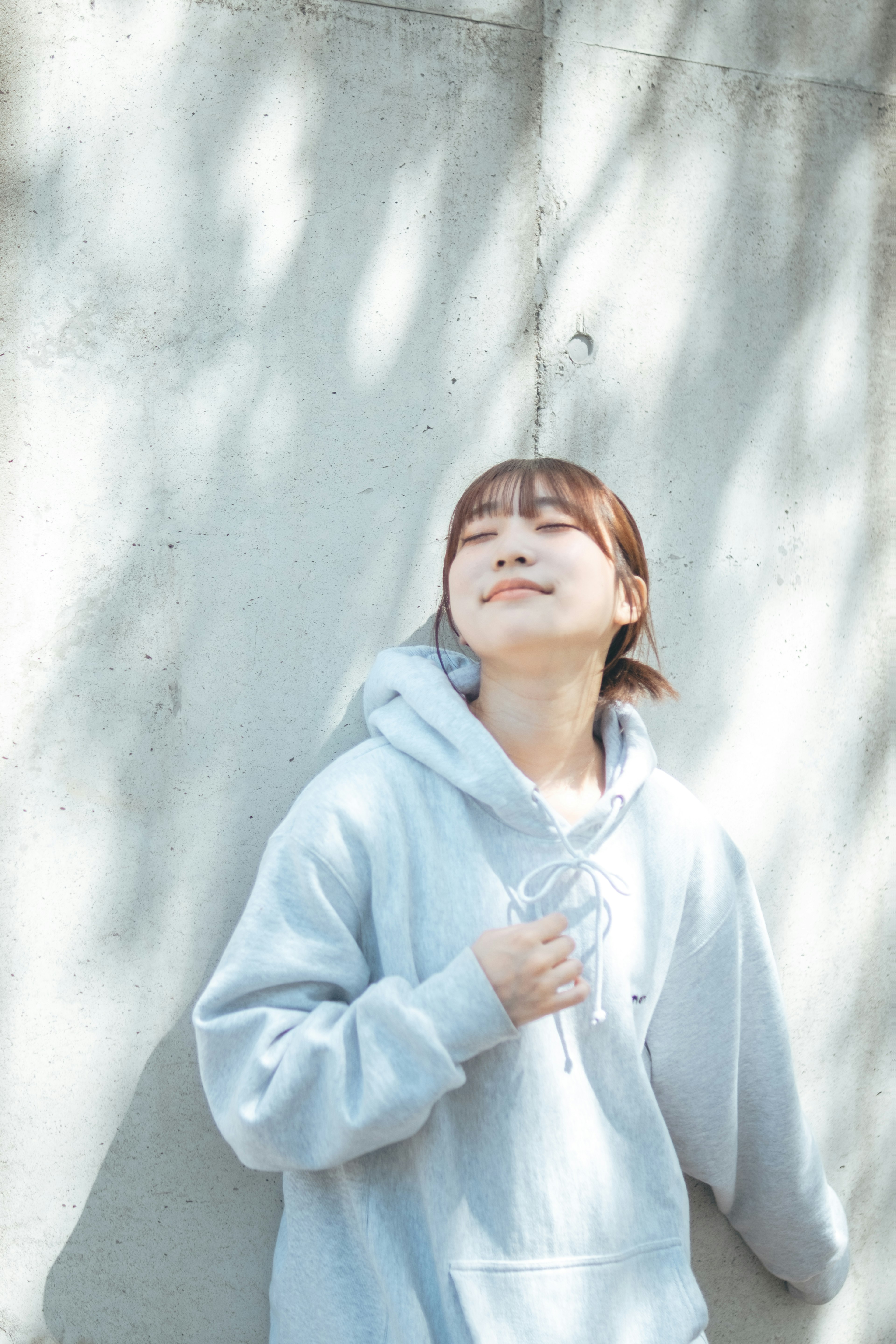 Young woman in a light blue hoodie leaning against a wall with a serene expression and closed eyes