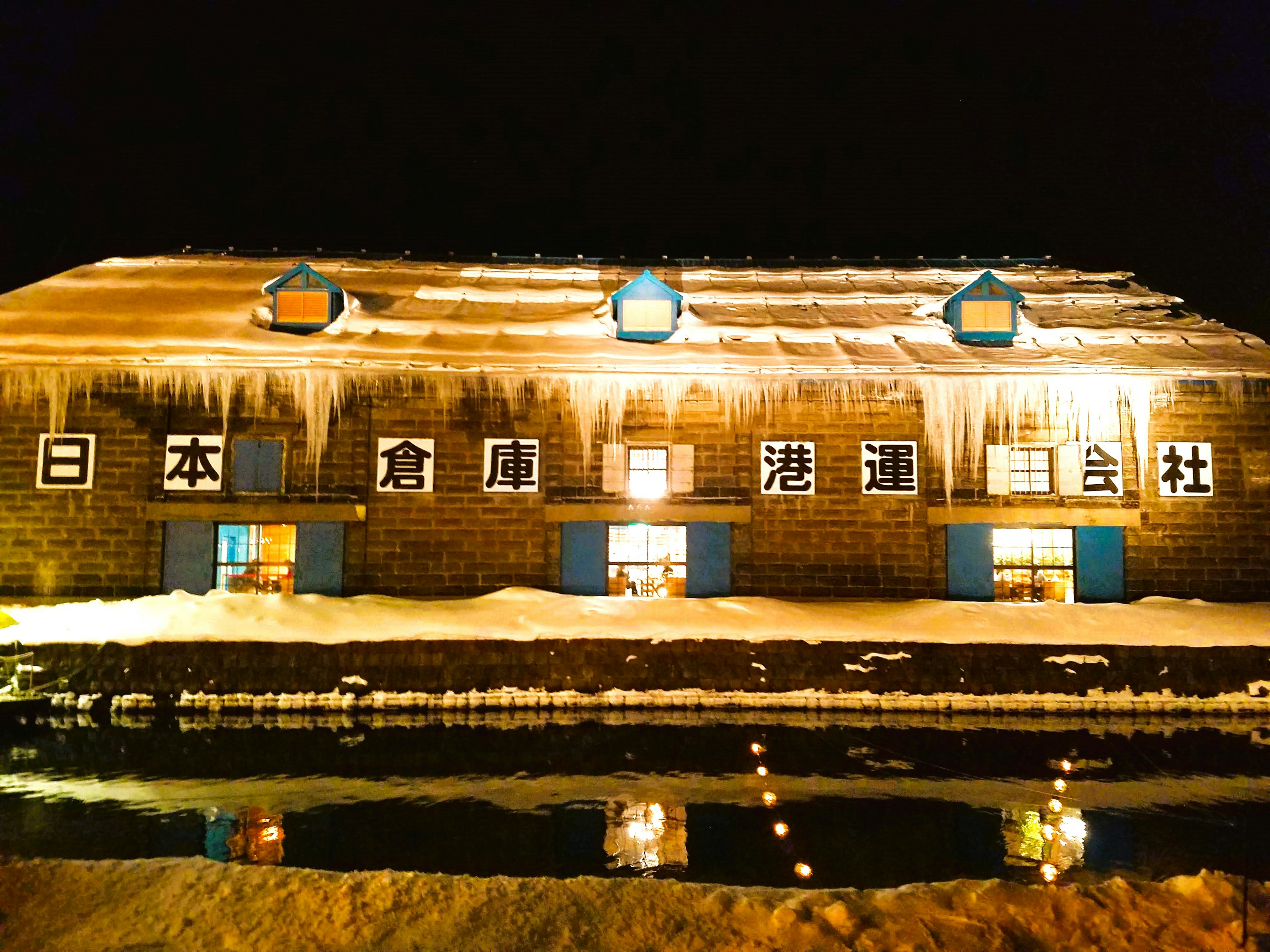 Außenansicht eines schneebedeckten Lagers bei Nacht mit blauen Fenstern und warmem Licht