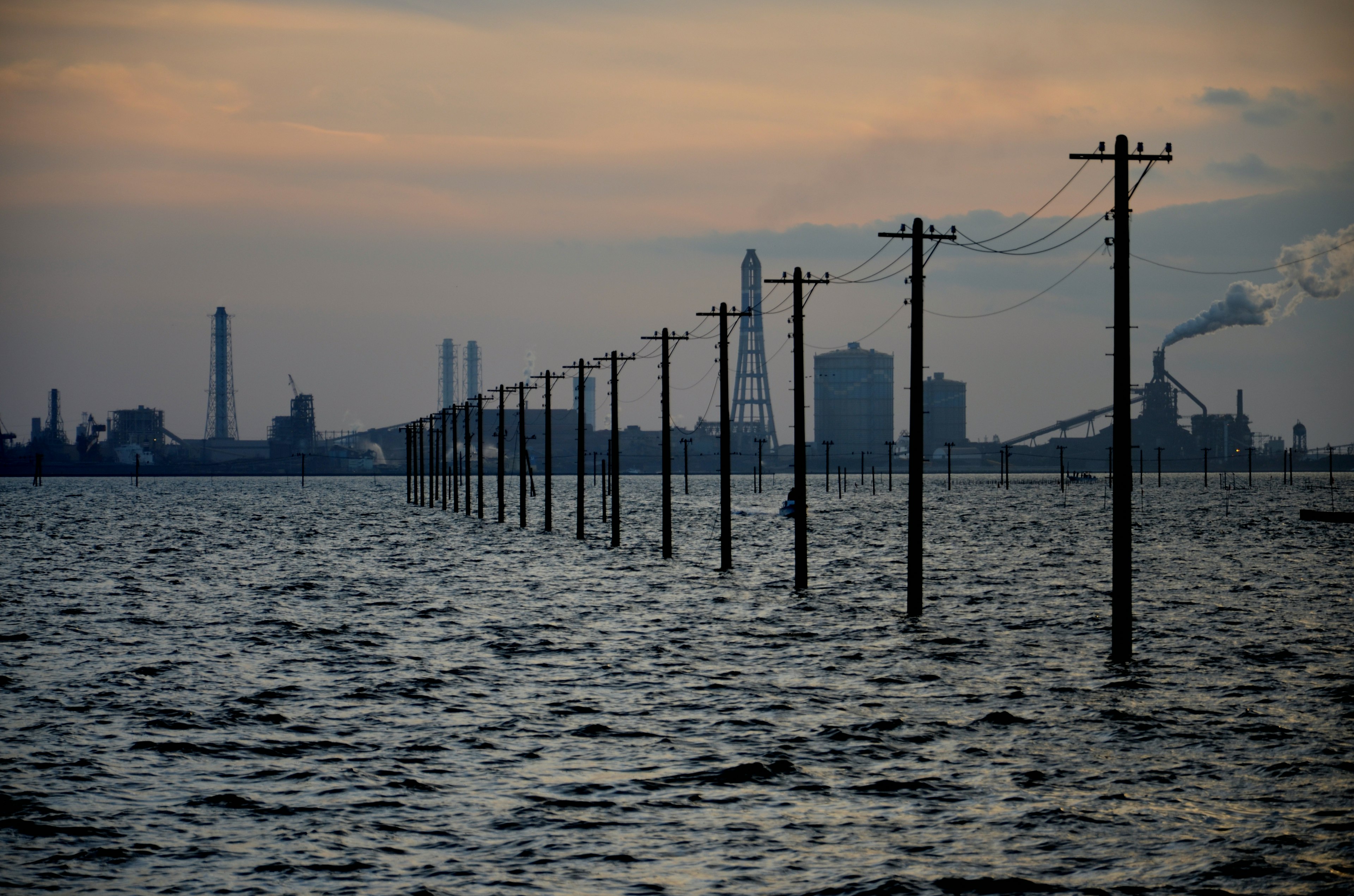 Stromleitungen, die aus dem Wasser ragen, mit einer Industrie-Skyline bei Sonnenuntergang