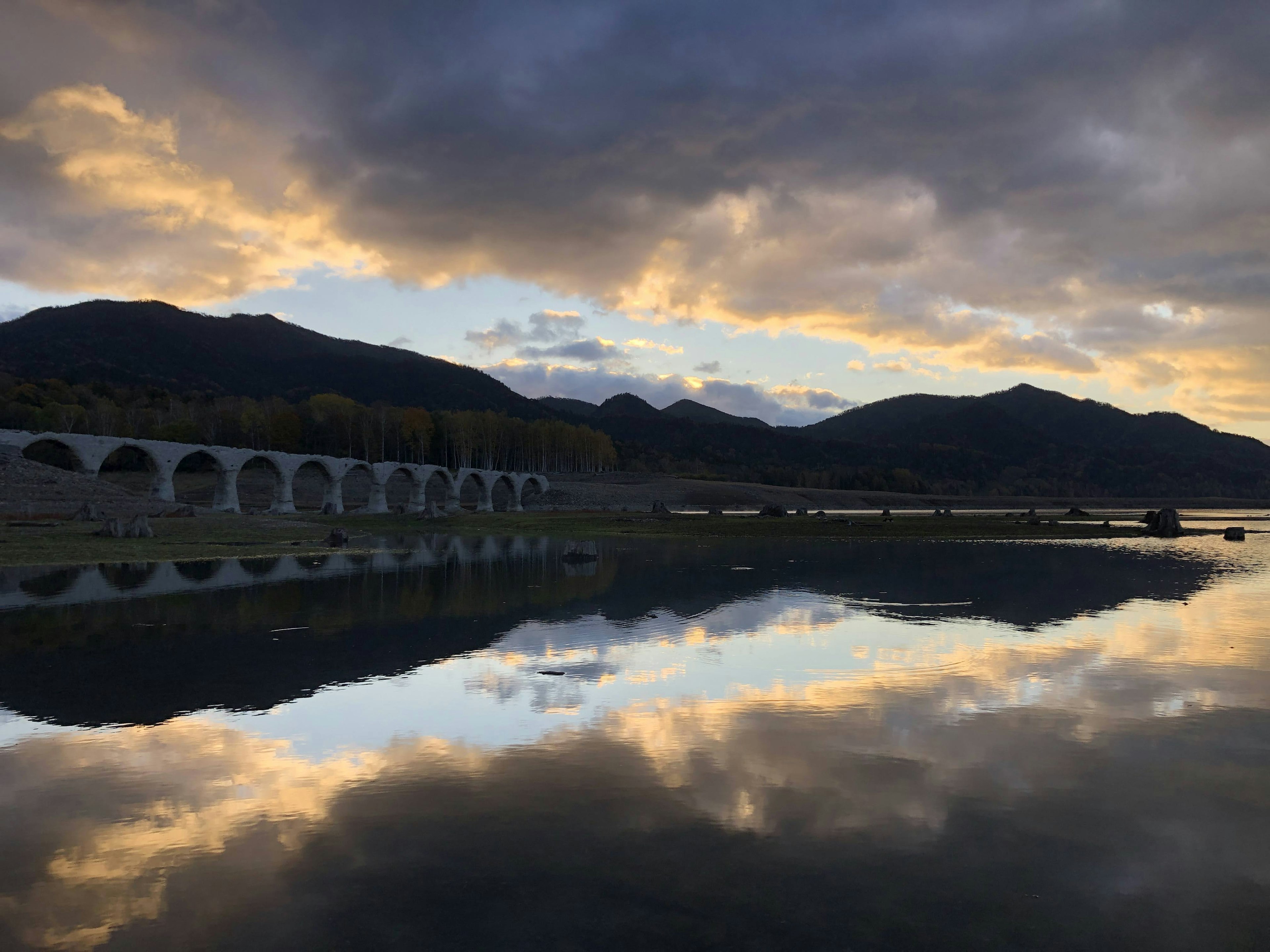 Vista escénica de un lago que refleja montañas y un colorido atardecer con un puente en arco