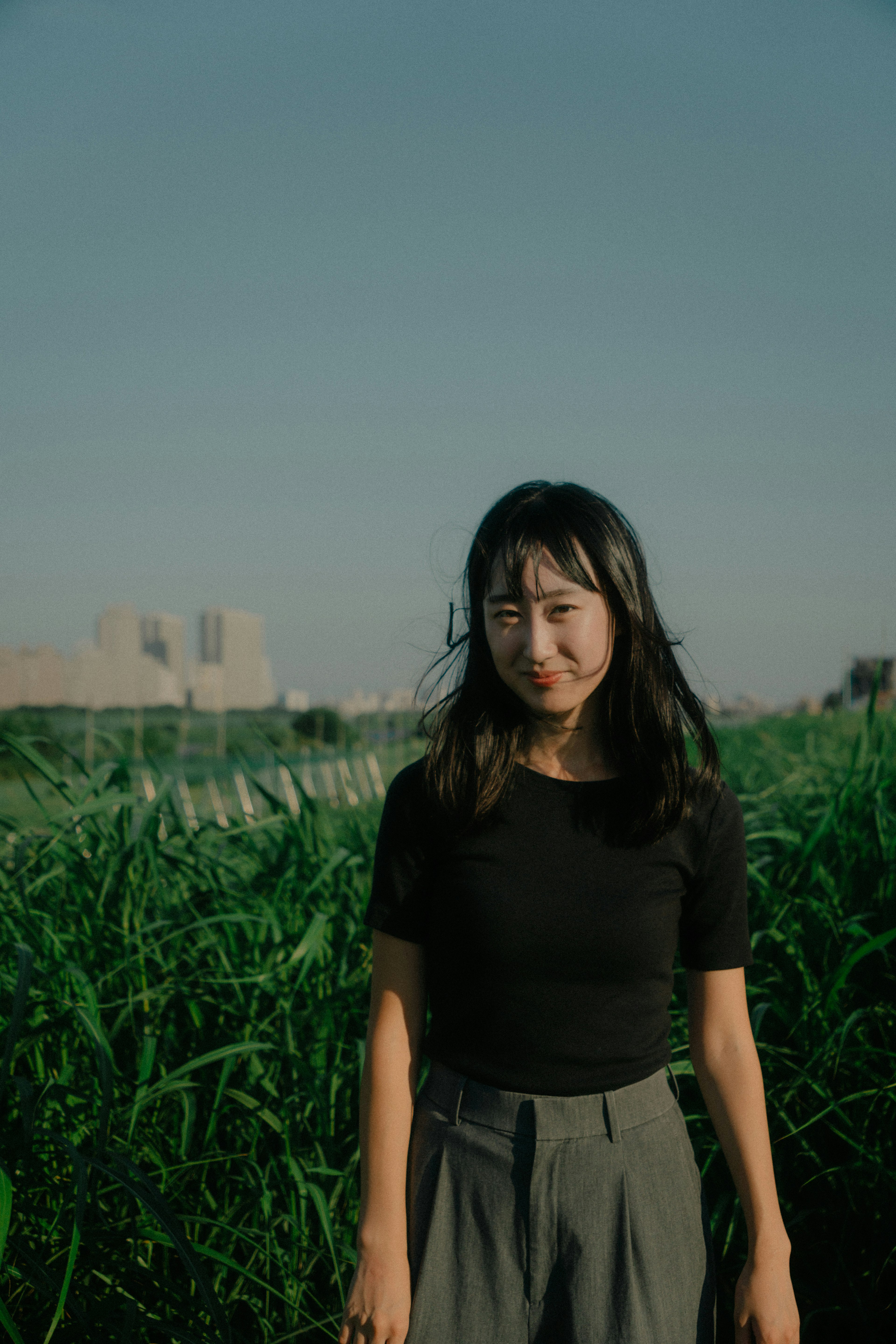 Woman standing under blue sky surrounded by green grass wearing a simple black top and gray pants