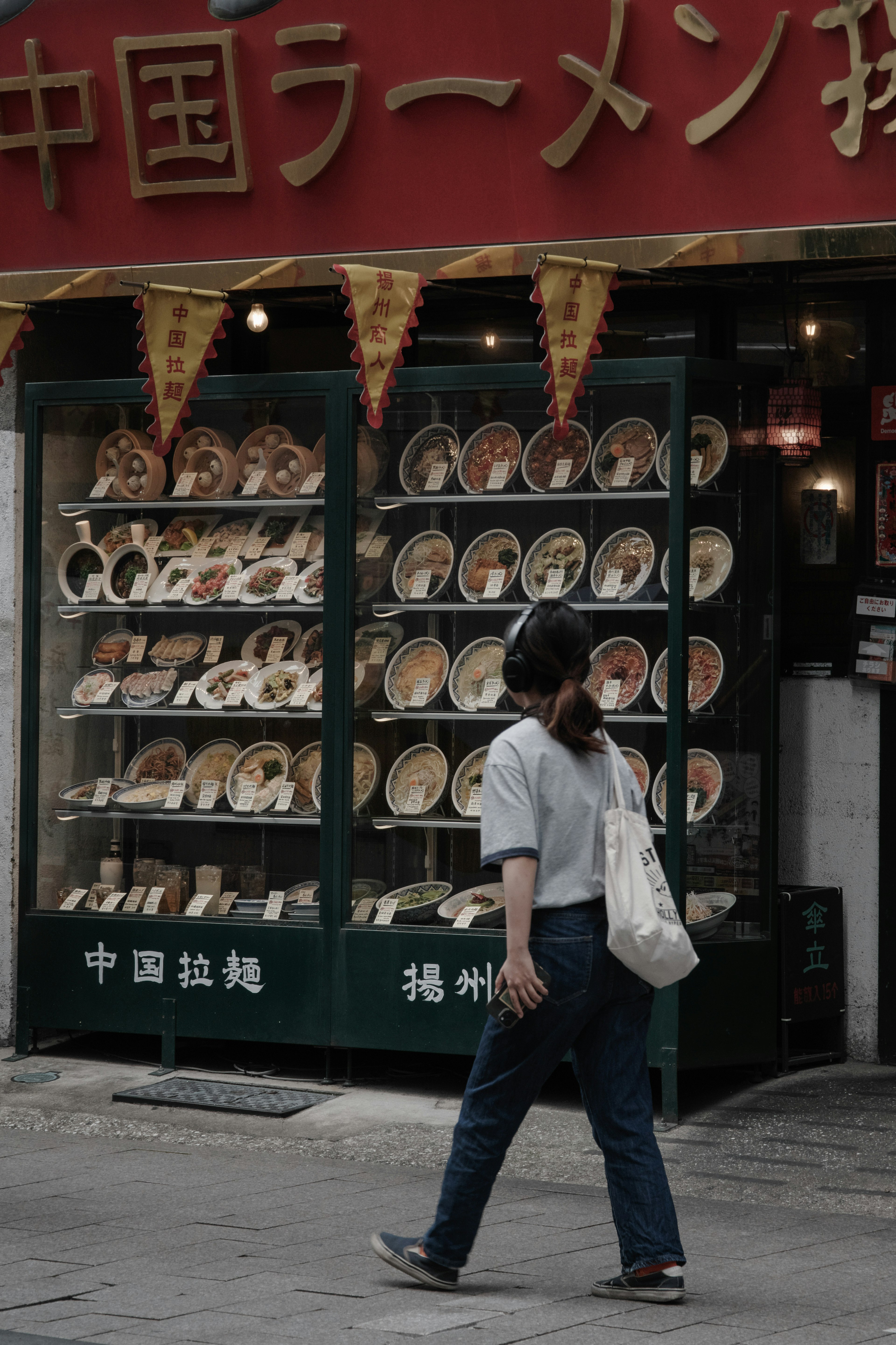 女性走在拉麵店前，展示櫃裡有許多盤子