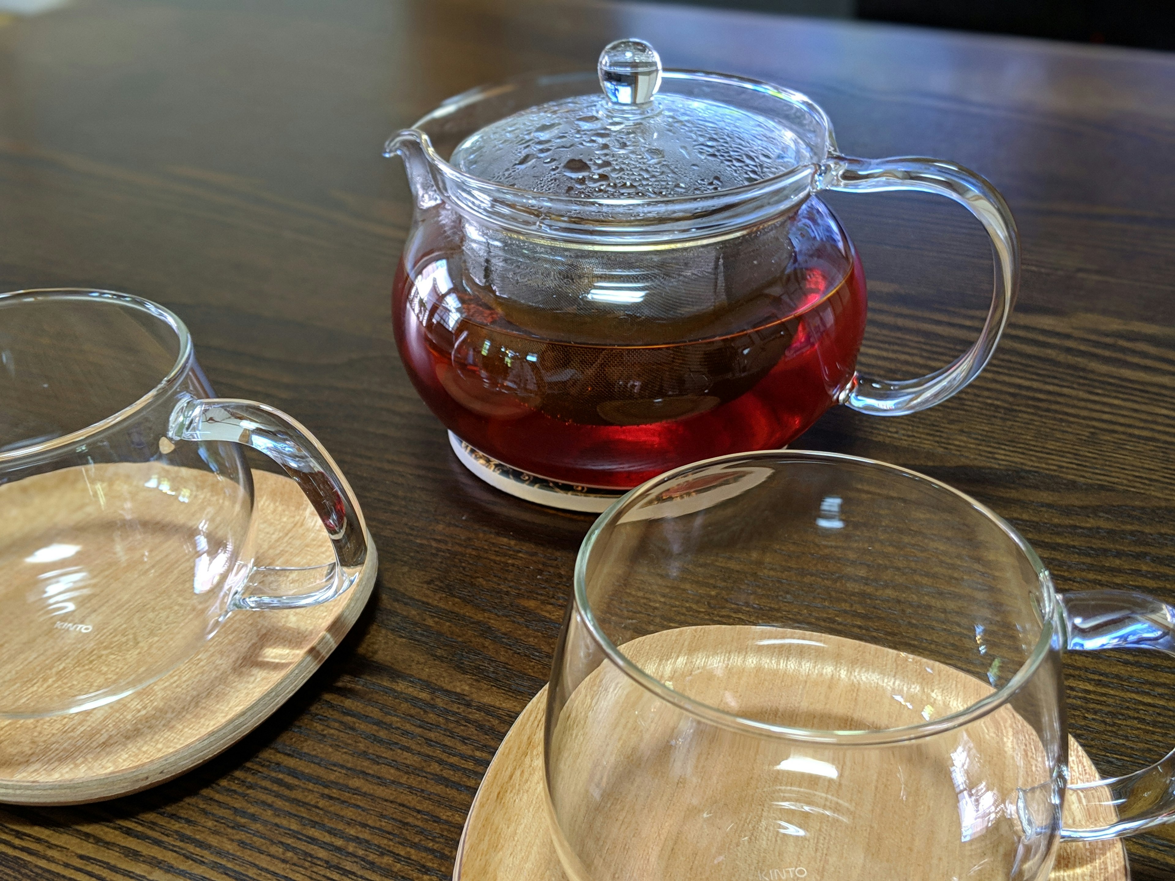 Une théière en verre remplie de thé rouge à côté de deux tasses en verre transparent sur une table en bois