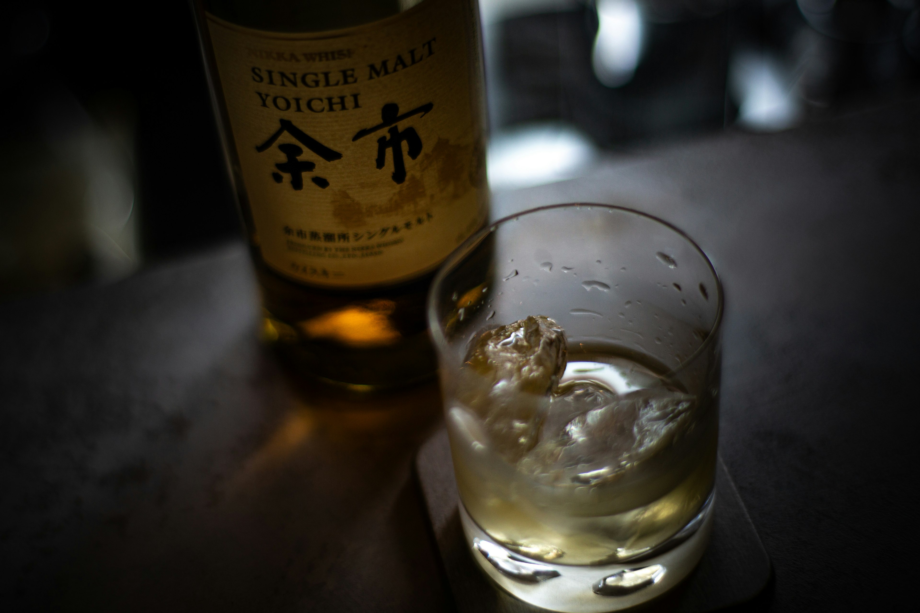 A whiskey glass with ice next to a bottle on a dark background