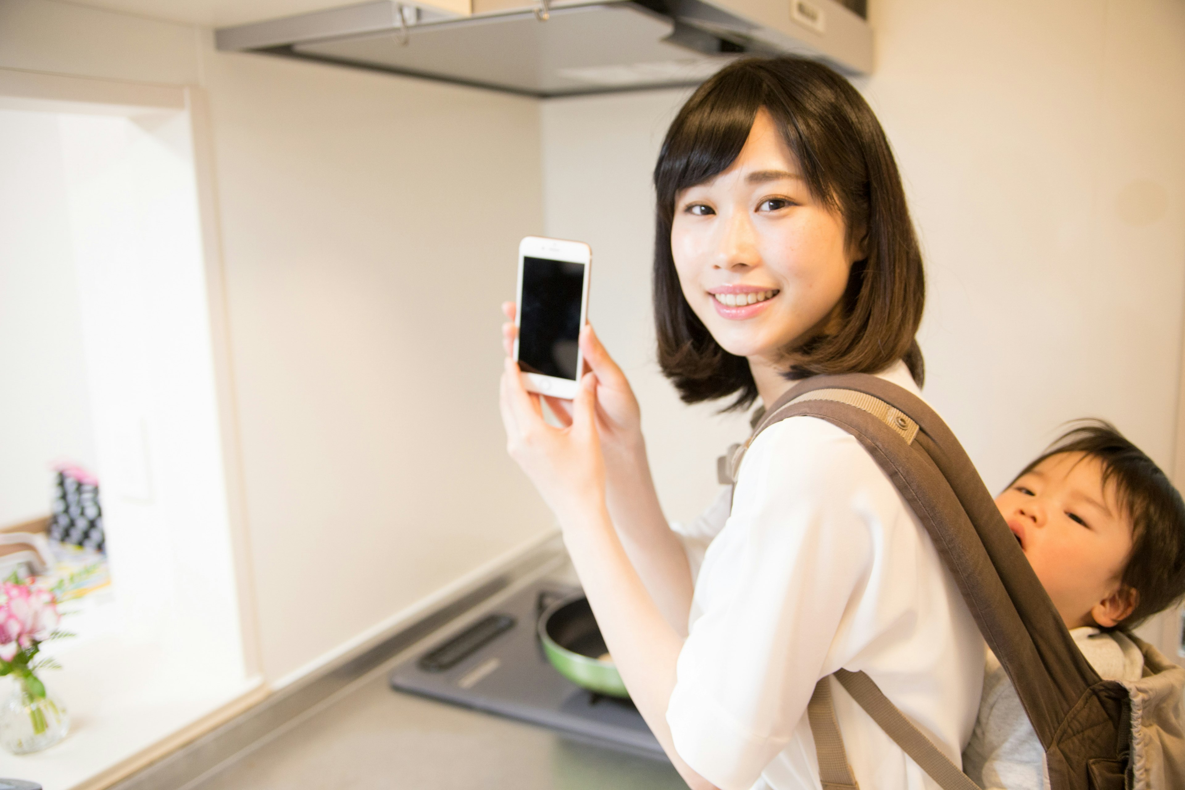 Mother holding a smartphone in the kitchen with her child on her back