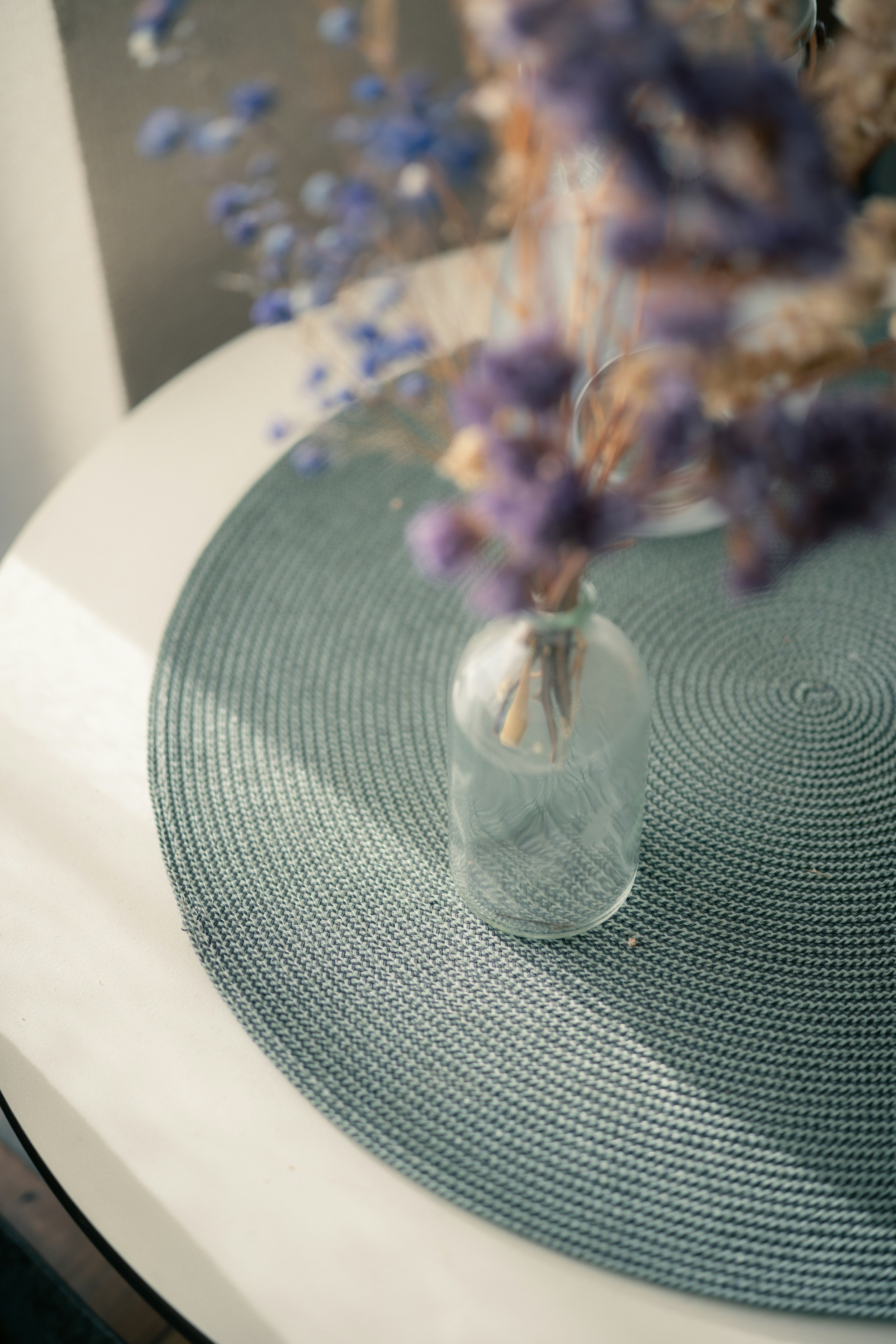 Un vase transparent avec des fleurs violettes posé sur un tapis bleu rond