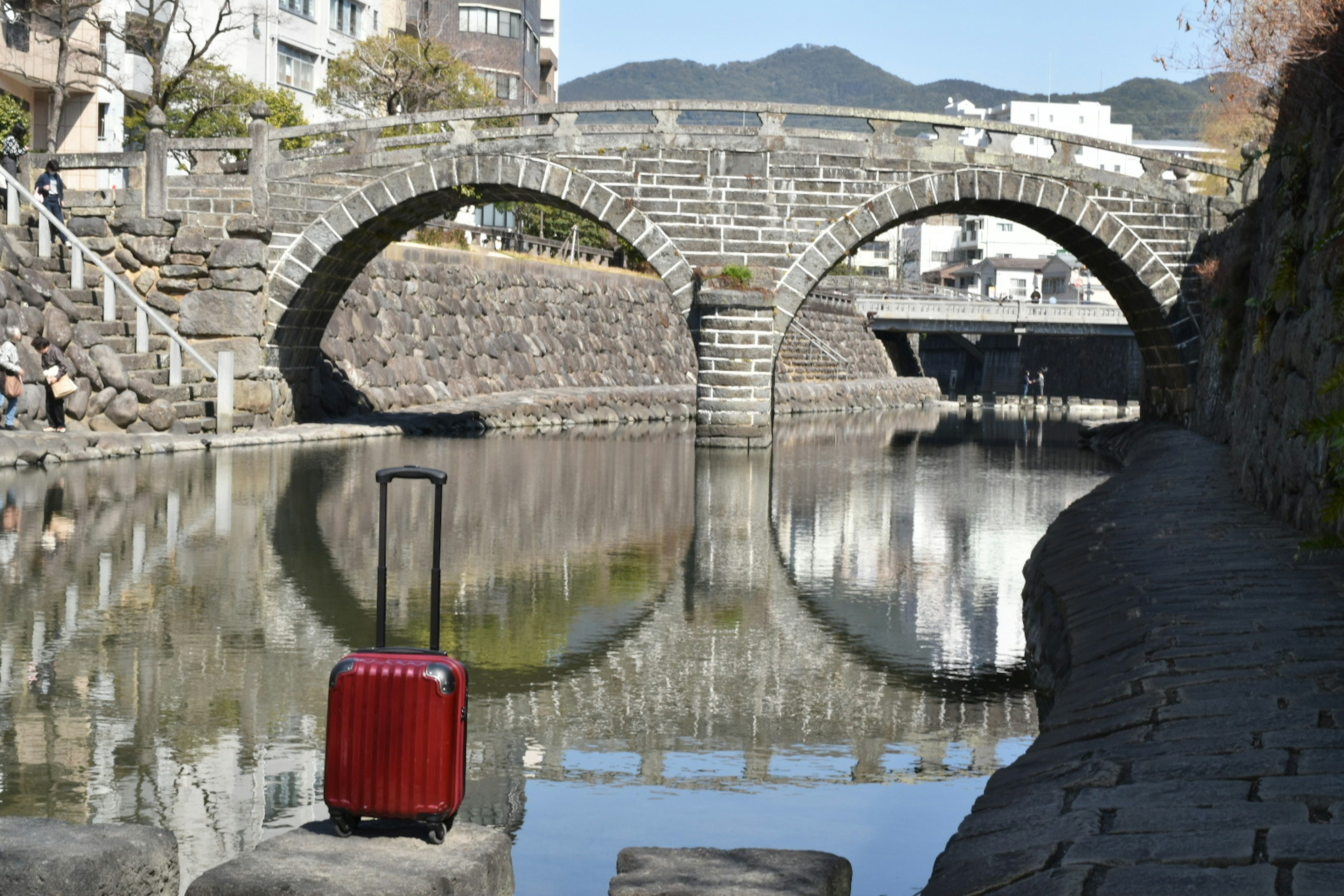 A red suitcase near a tranquil waterway with stone bridges reflecting in the water