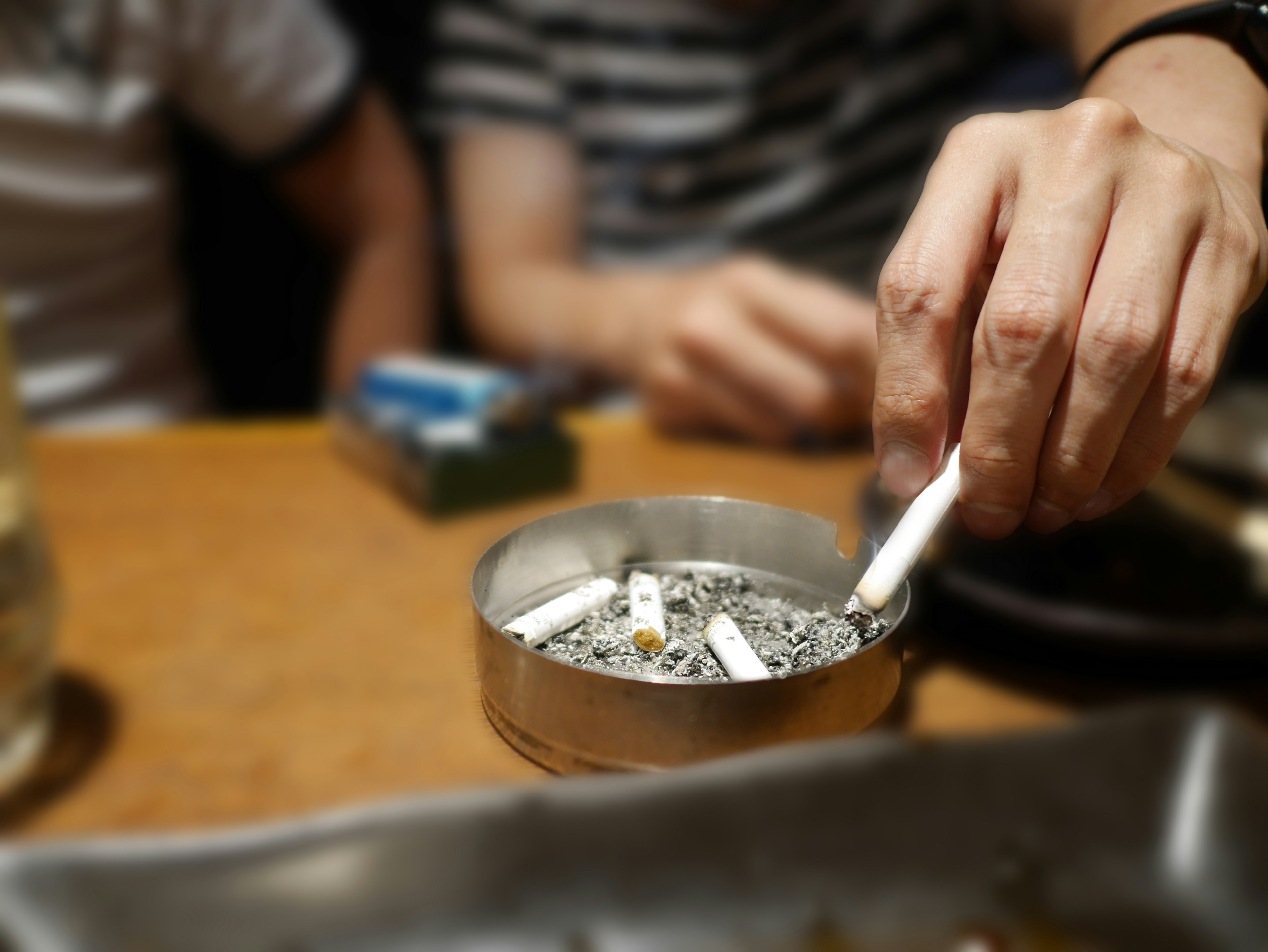 Hand hält eine Zigarette über einem Aschenbecher mit Zigaretten und unscharfem Hintergrund