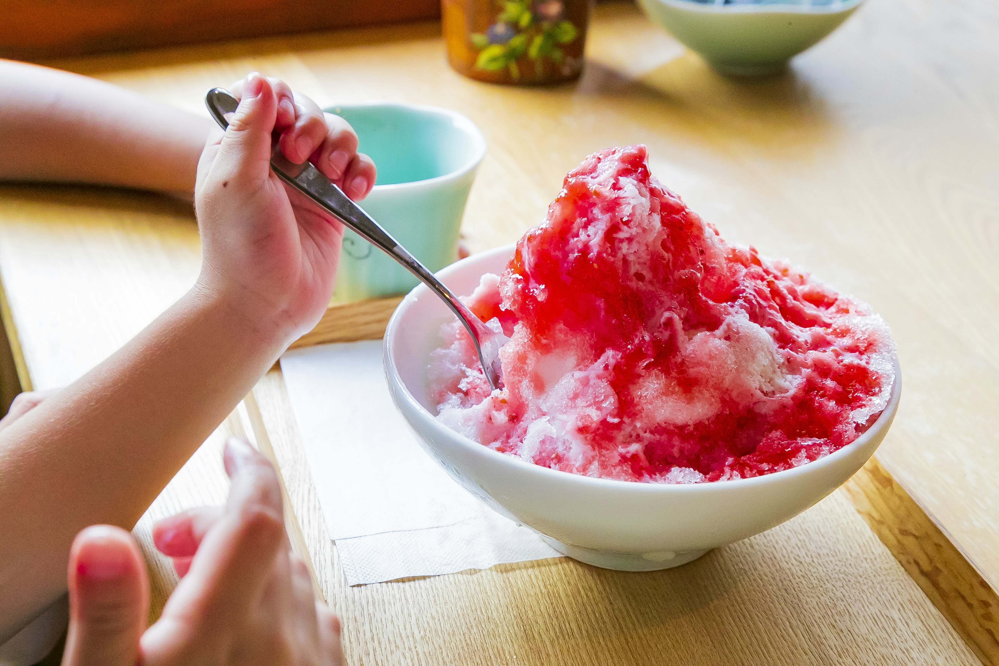 Hand hält einen Löffel über eine Schüssel mit rotem Sirup-bedecktem Shaved Ice