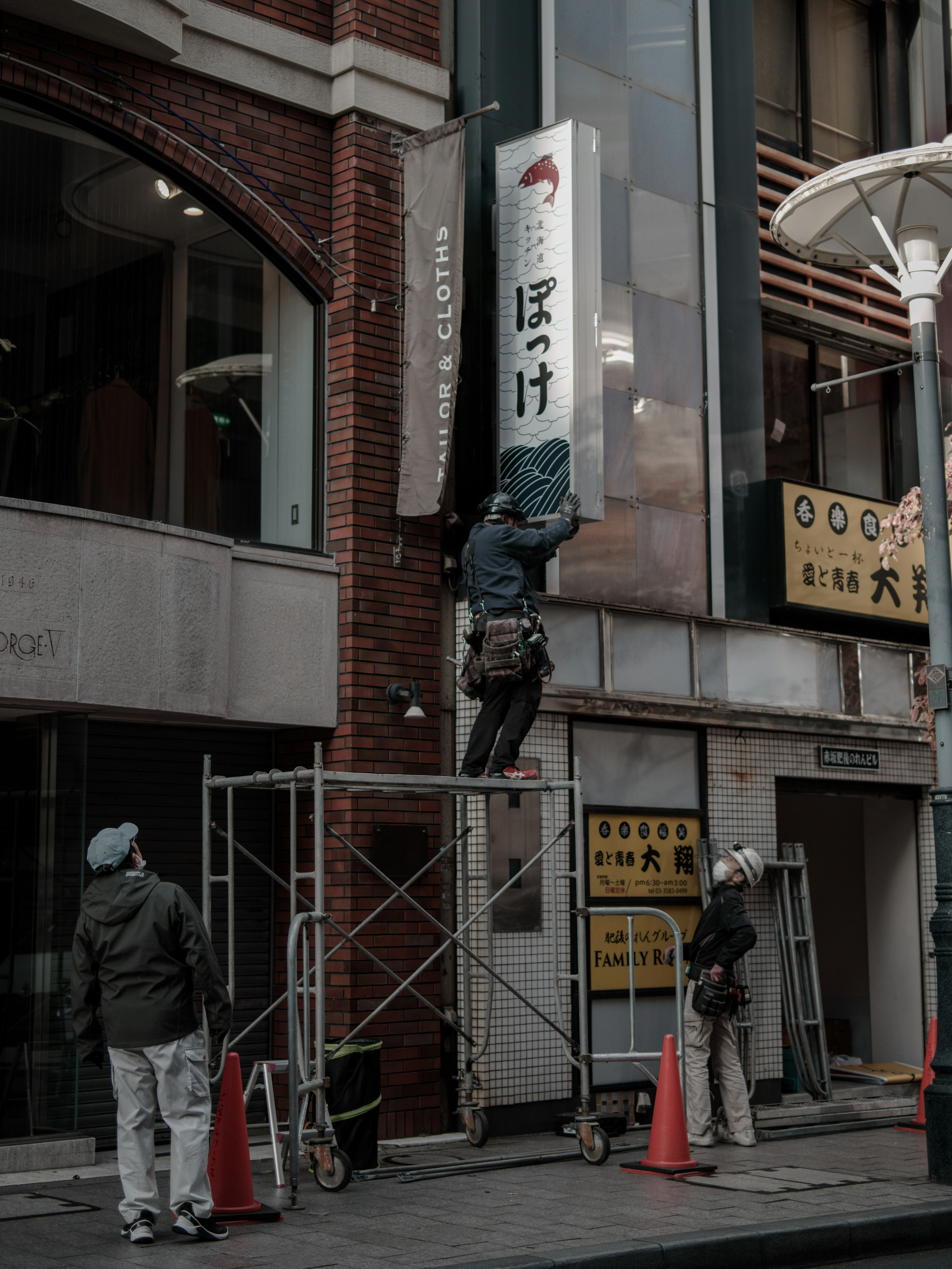 工事中の看板を取り付ける作業員と足場が見える街の風景