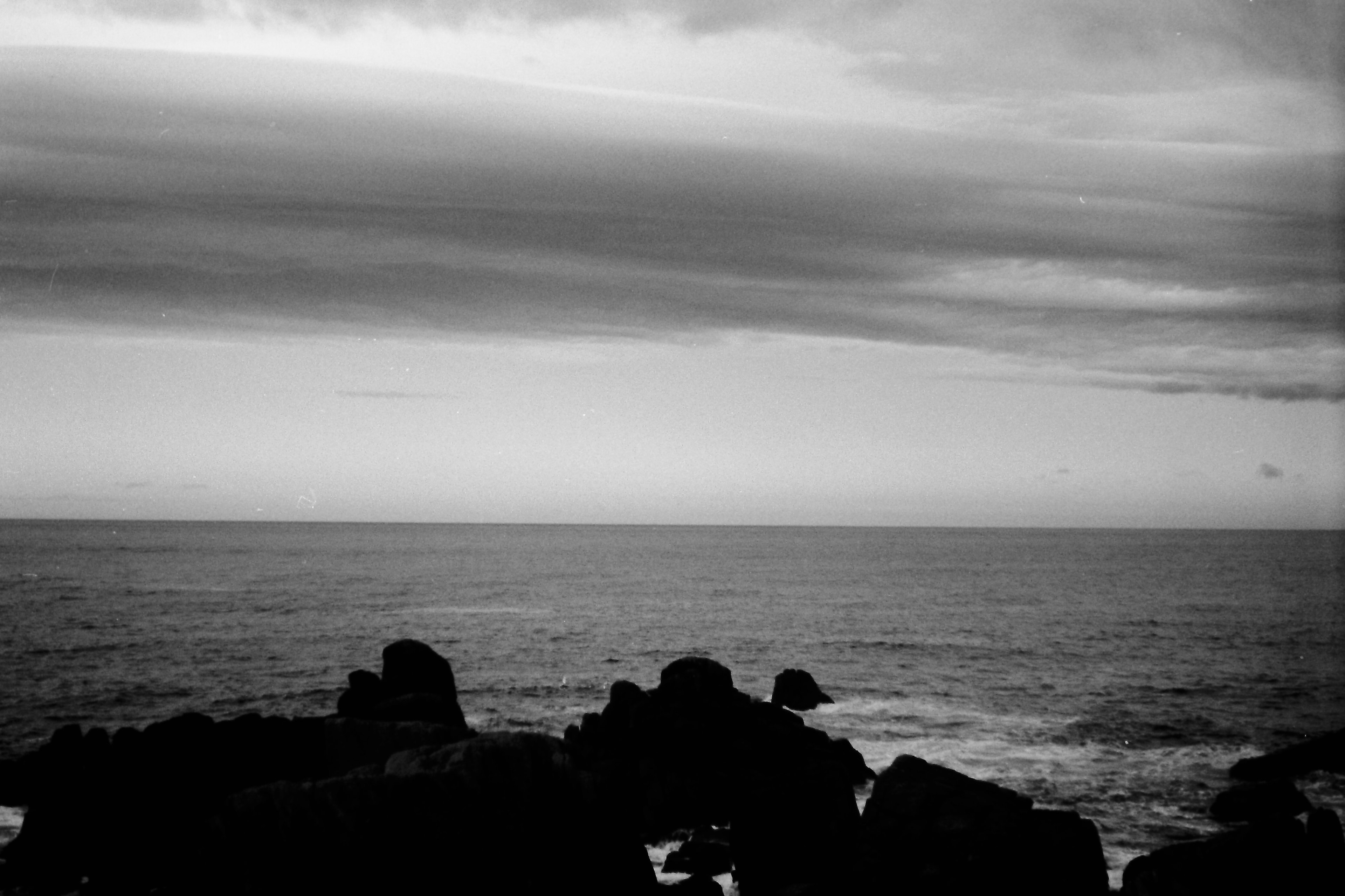 Paisaje marino en blanco y negro con rocas y agua tranquila bajo un cielo nublado